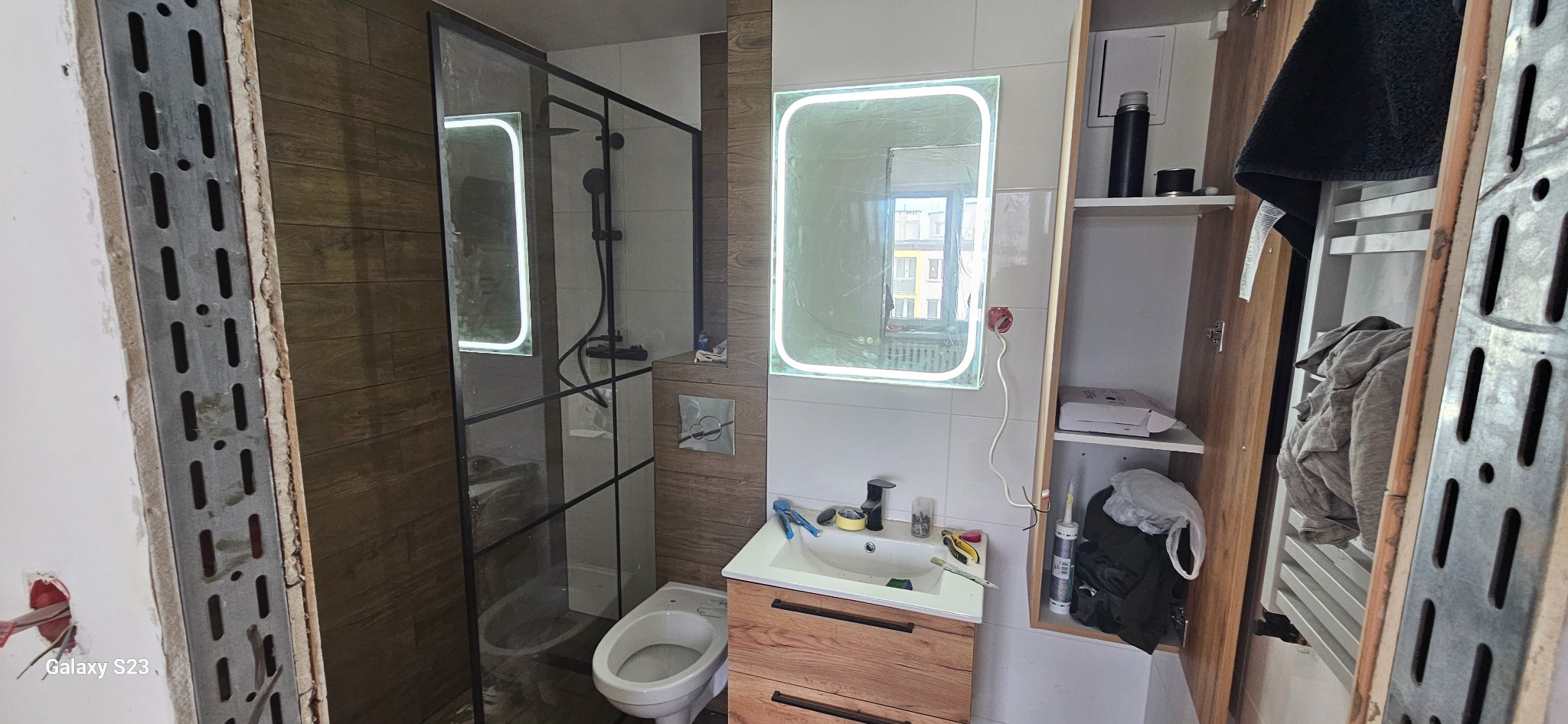 Łazienka w trakcie prac wykończeniowych: widoczna kabina prysznicowa z czarnymi ramami, umywalka z drewnianą szafką, lustro z podświetleniem LED w folii ochronnej, bidet, oraz otwarty regał...