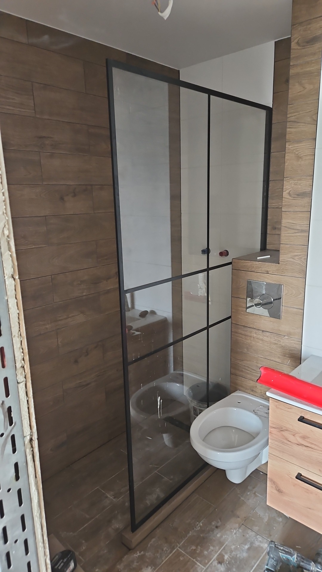 Montaż kabiny prysznicowej z czarnymi profilami w łazience z płytkami imitującymi drewno, widoczne elementy instalacji i narzędzia.