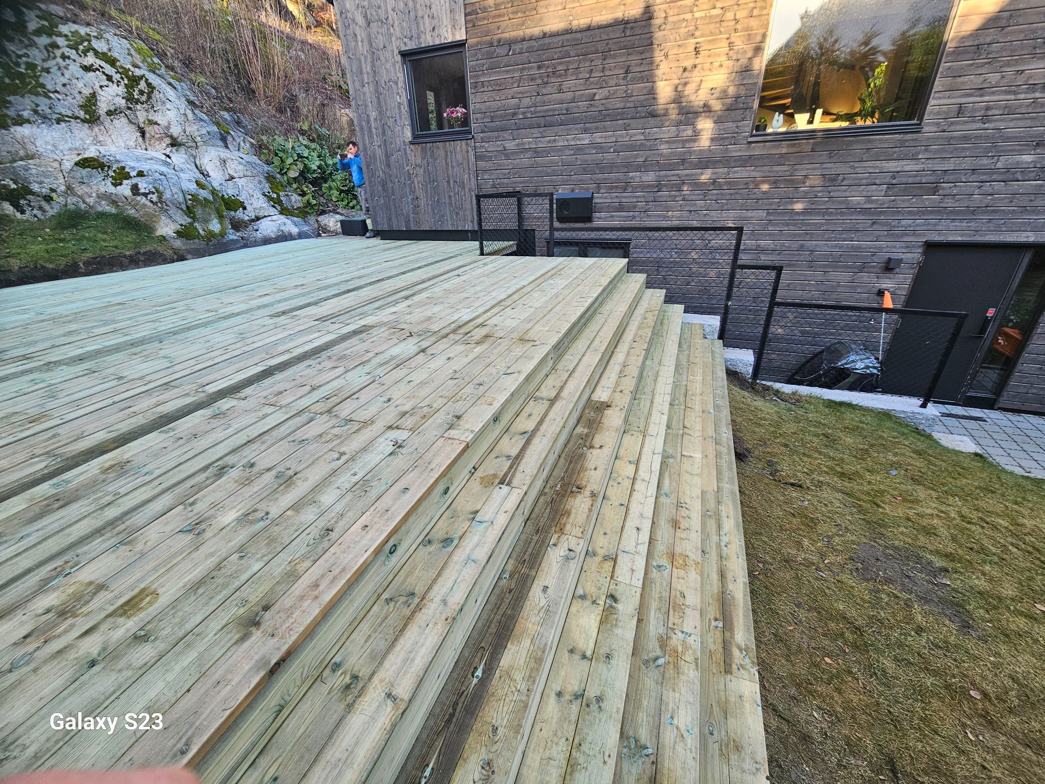Świeżo zbudowany taras drewniany z widocznymi schodami, przylegający do ciemnej, drewnianej elewacji domu, z fragmentem skały porośniętej mchem w tle. Mężczyzna w niebieskiej koszuli obserwuje z boku.