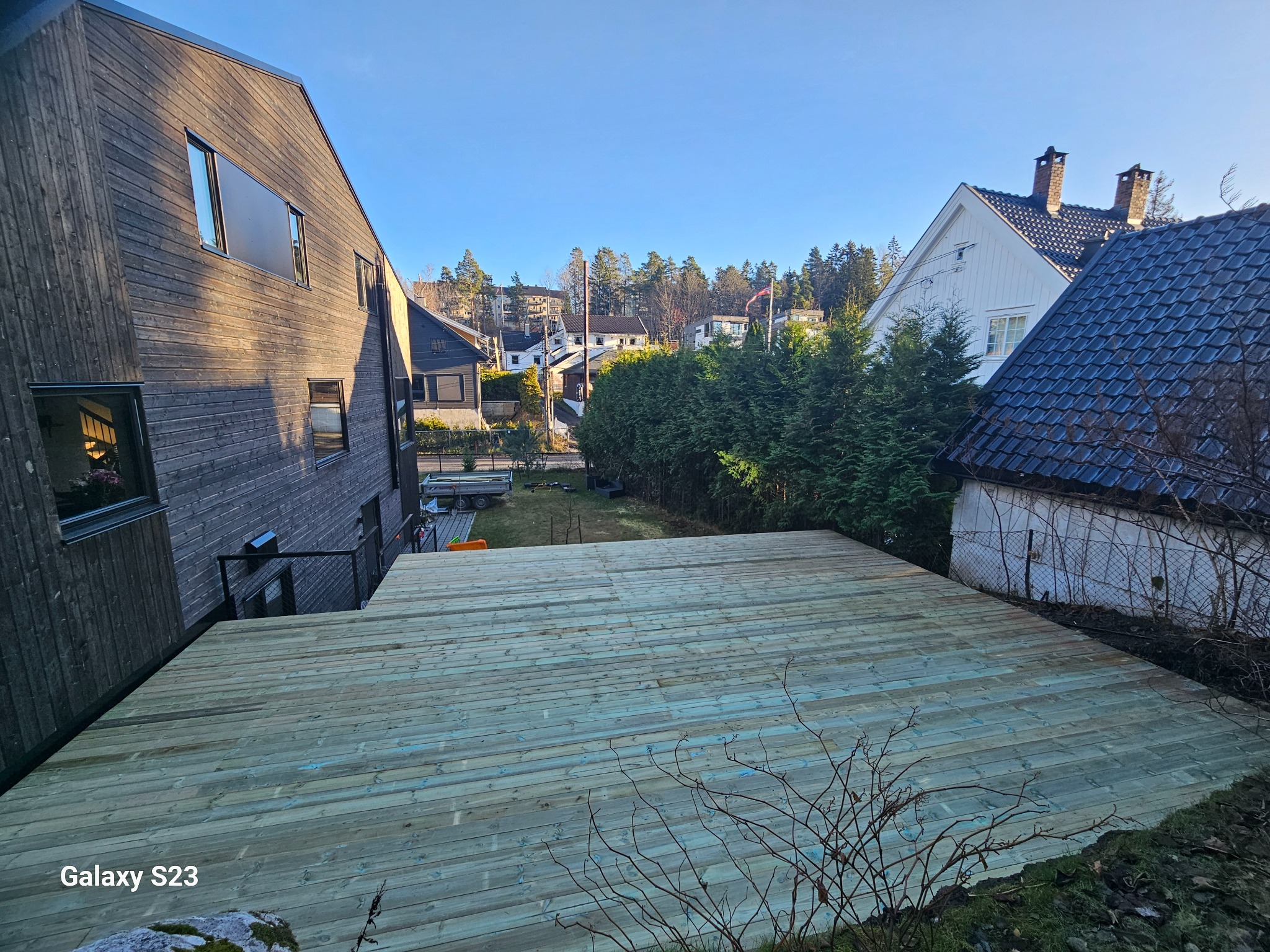 Widok z góry na nowo wybudowany taras drewniany z desek w odcieniach zieleni i szarości, z widocznymi domami w tle i fragmentem skalnego podłoża w dolnej części kadru.