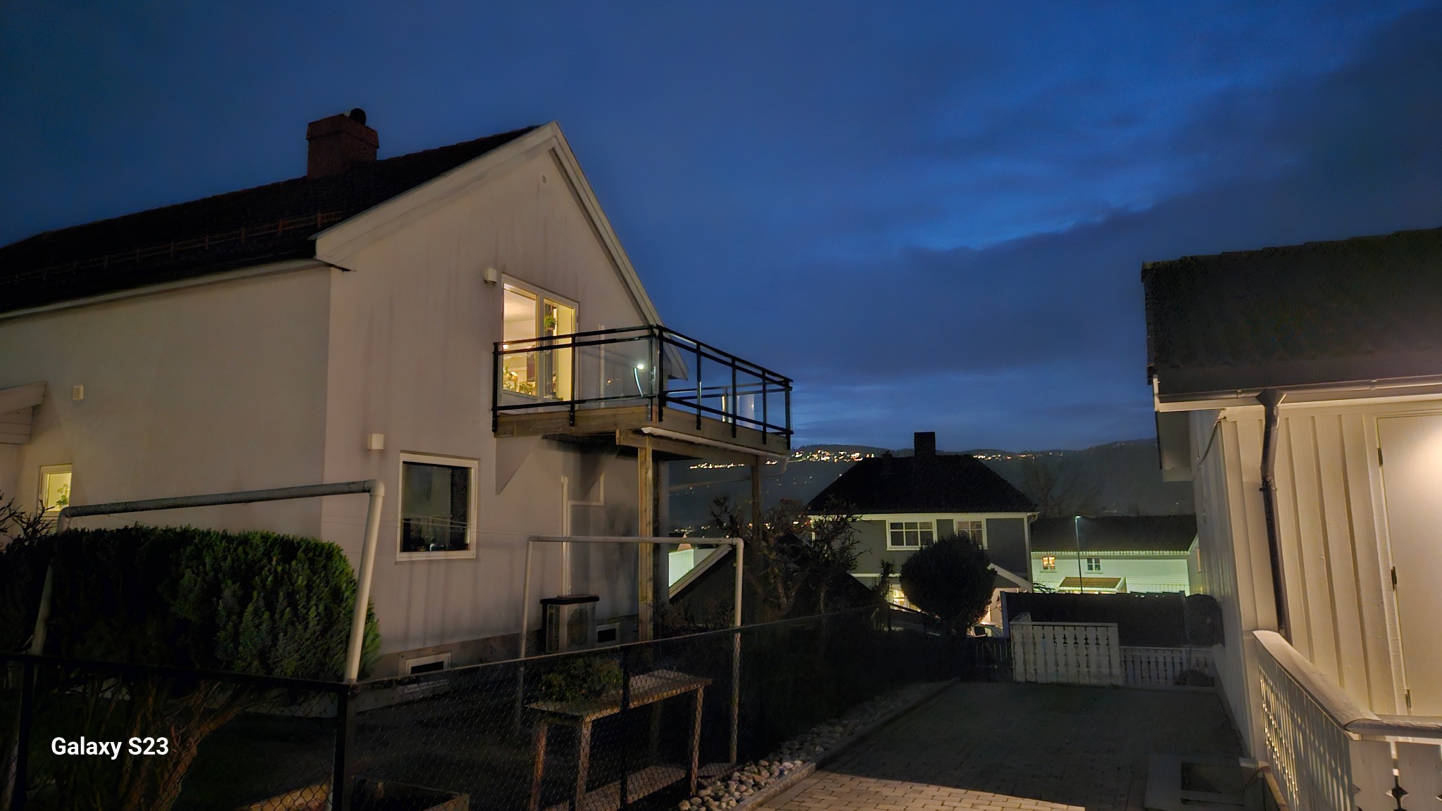 Dom z drewnianym tarasem z czarną balustradą i szklanym wypełnieniem, widok na oświetlone miasto w oddali pod wieczornym niebem.