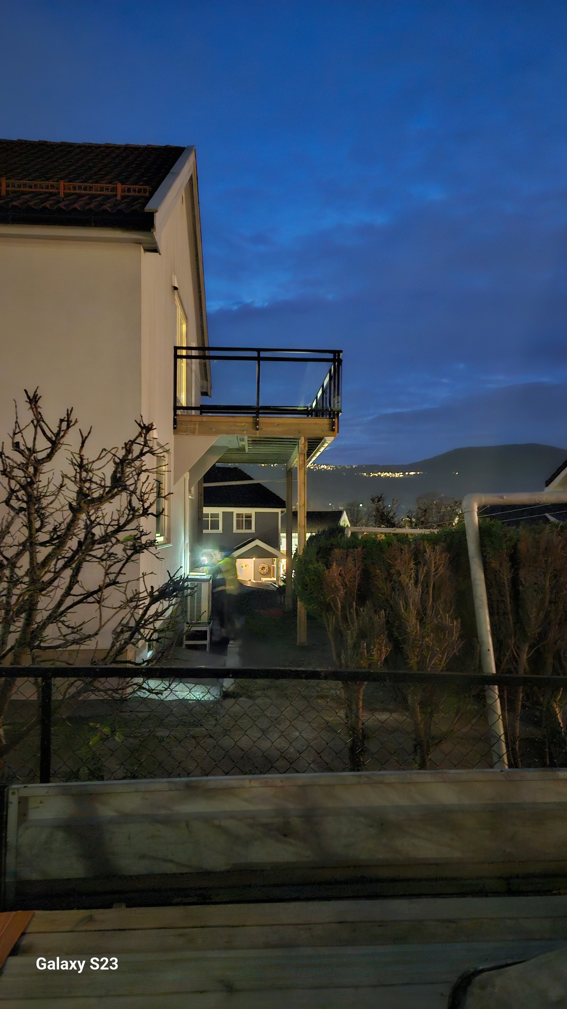 Nocne ujęcie domu z drewnianym balkonem z czarną balustradą, widok zza ogrodzenia, w tle zabudowania i oświetlone miasto na wzgórzu pod błękitnym niebem.