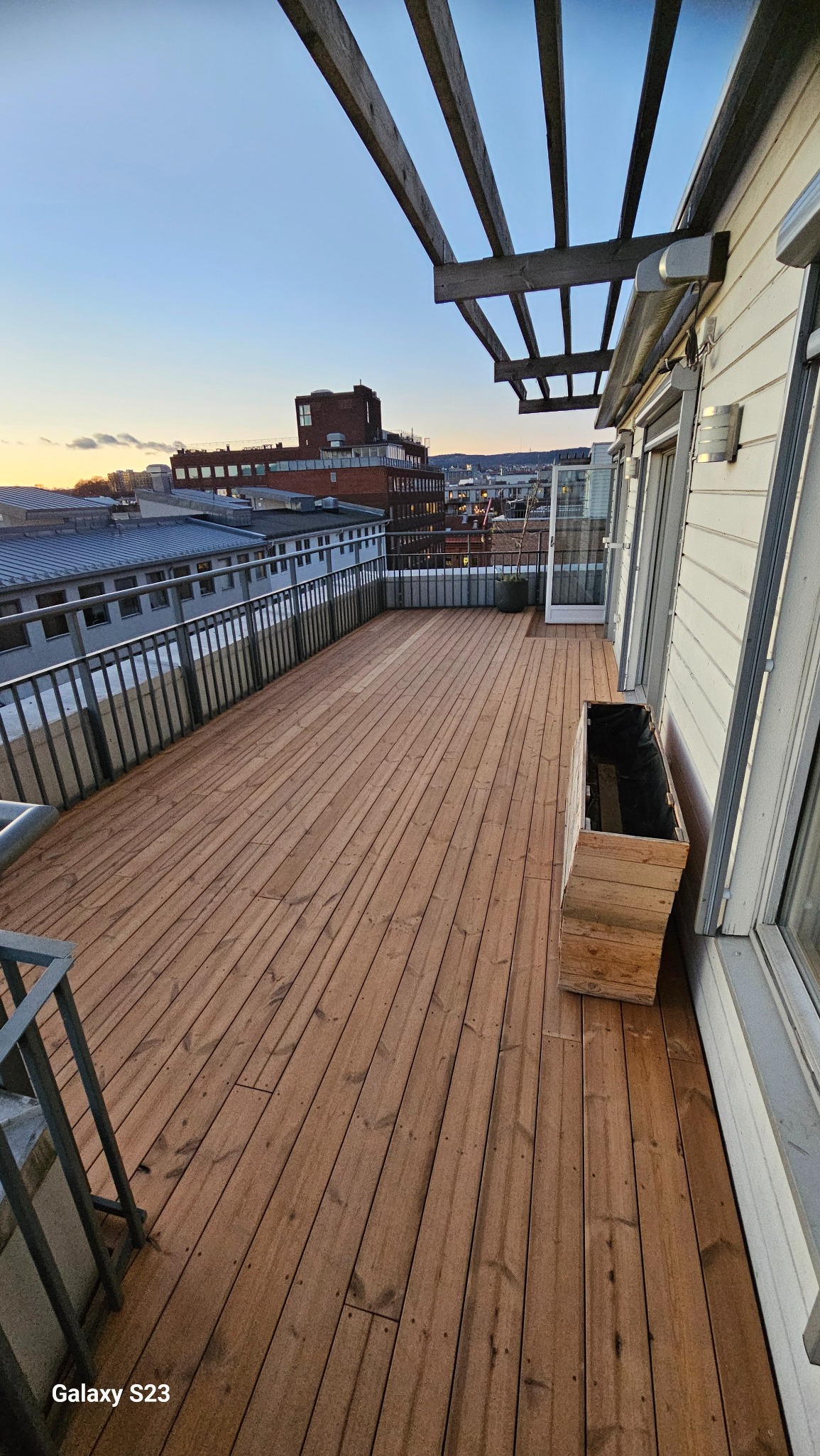 Jasny taras drewniany z widokiem na miasto o zmierzchu, drewniana skrzynia na rośliny przy ścianie budynku.