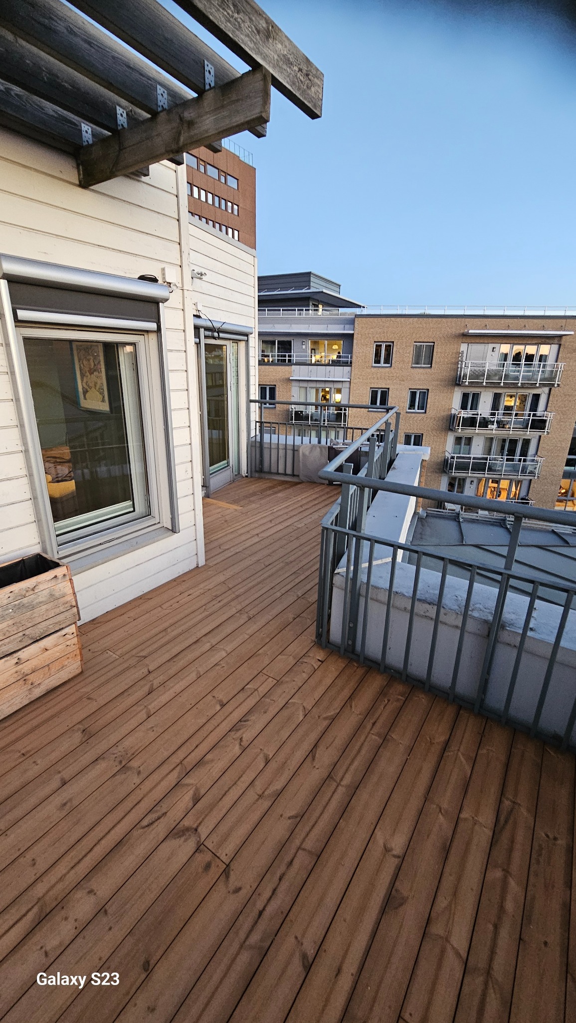 Widok z góry na drewniany taras z metalową barierką, z widokiem na sąsiedni budynek mieszkalny i błękitne niebo w tle. Na tarasie stoi drewniana skrzynka.