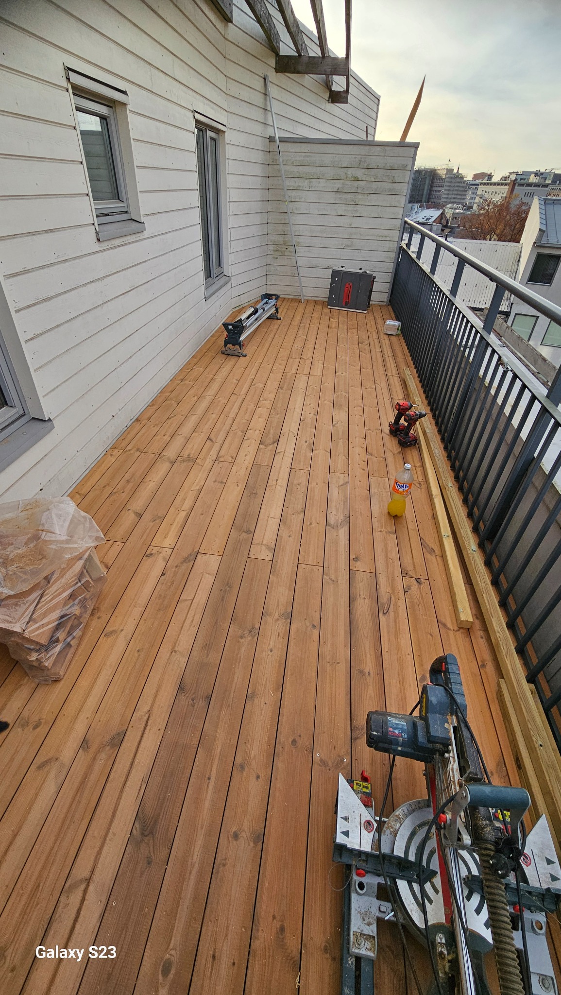 Montaż drewnianego tarasu na balkonie, widoczne deski, piła ukośnica, wkrętarka, poziomica i butelka napoju.