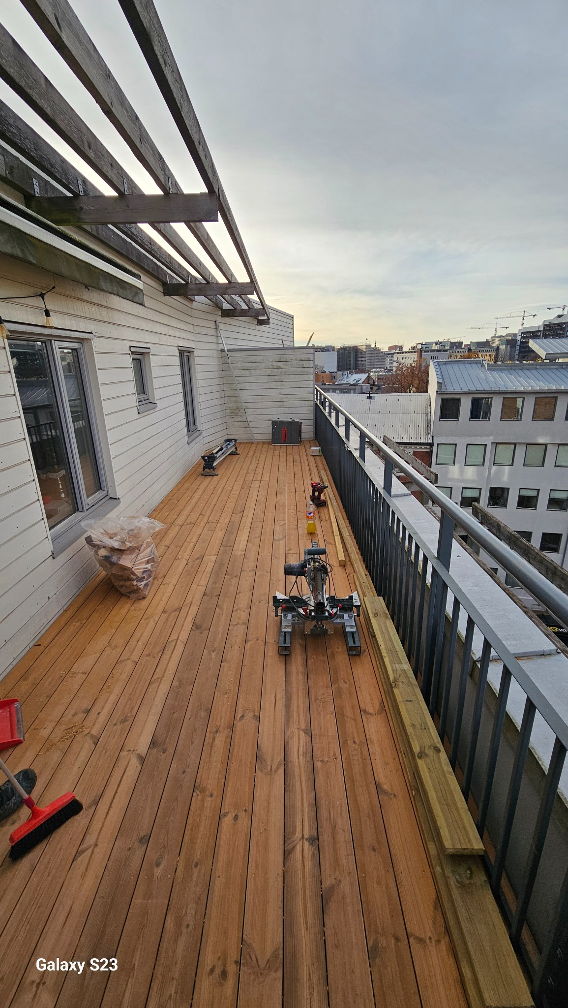 Montaż desek na tarasie drewnianym na dachu budynku, widoczna piła ukośnica, torba z materiałami, szczotka i panorama miasta w tle.