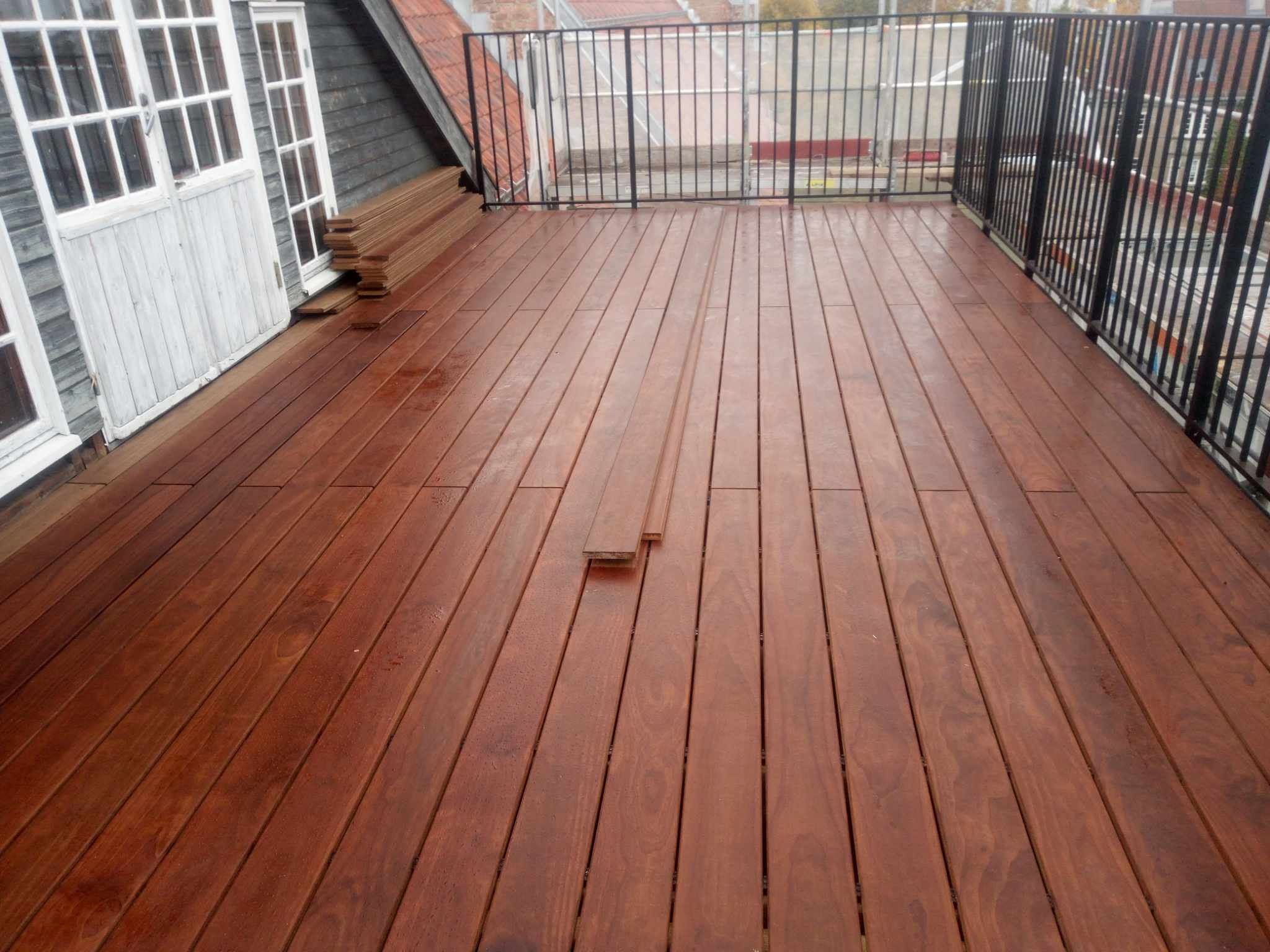 Montaż desek na tarasie drewnianym, widoczne ułożone deski i stos desek przy ścianie, czarna metalowa balustrada w tle.