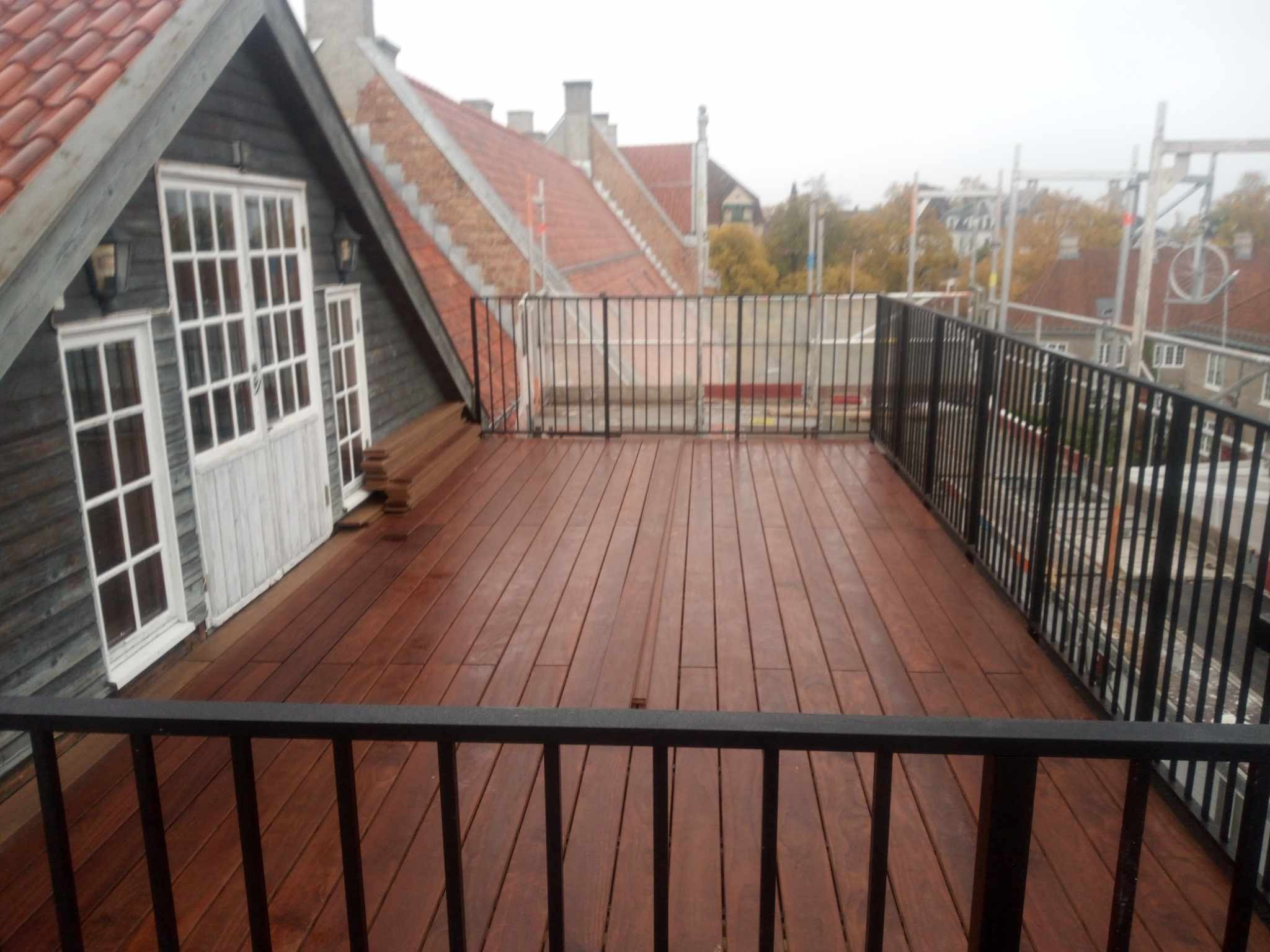 Nowy taras drewniany na dachu budynku z czarną metalową balustradą, widok na okoliczne dachy i rusztowania w tle.
