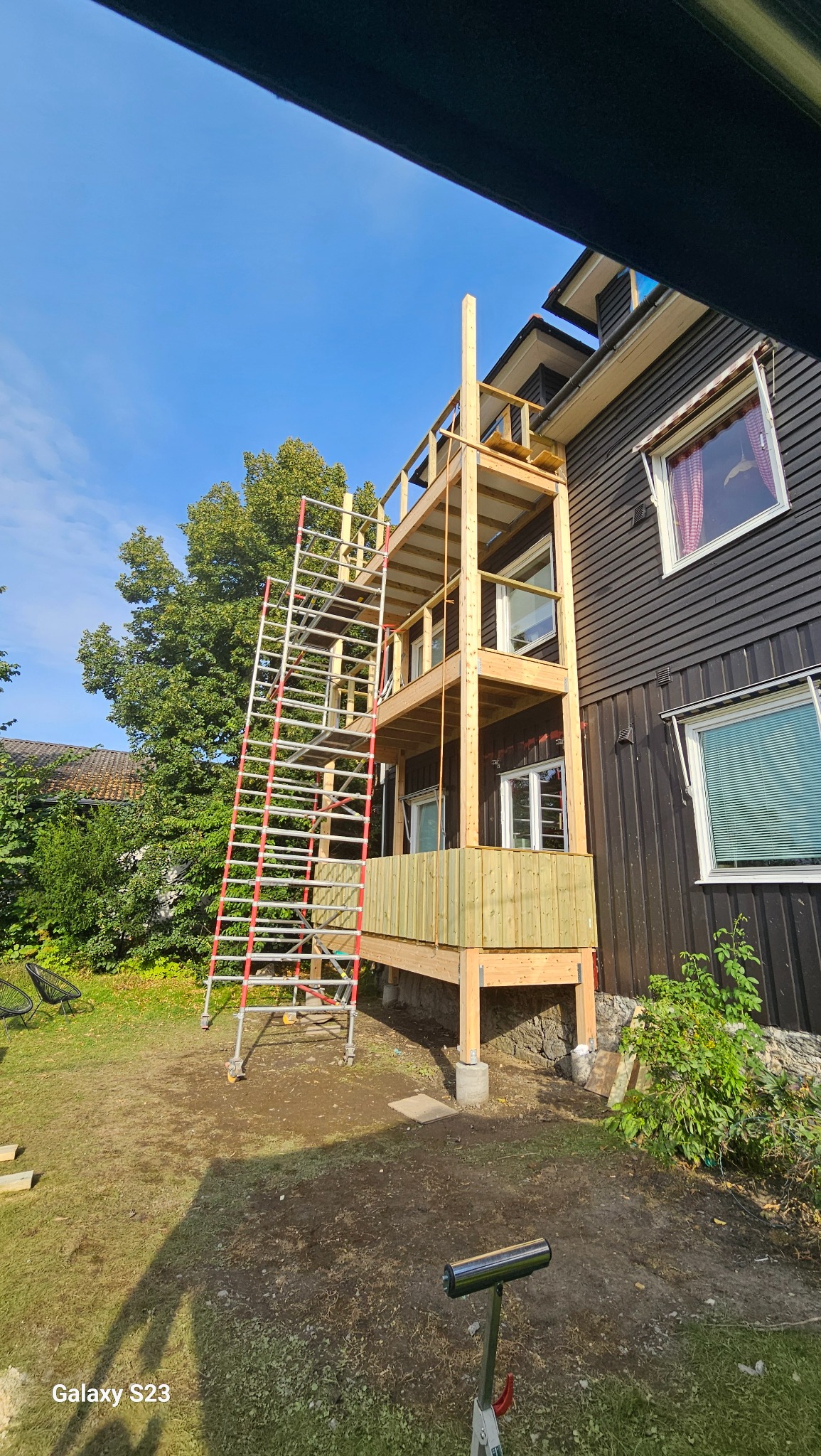 Wielopoziomowa konstrukcja tarasu drewnianego w trakcie budowy, z widocznymi rusztowaniami, surowym drewnem i elementami mocującymi, przy ciemnej elewacji domu z oknami.