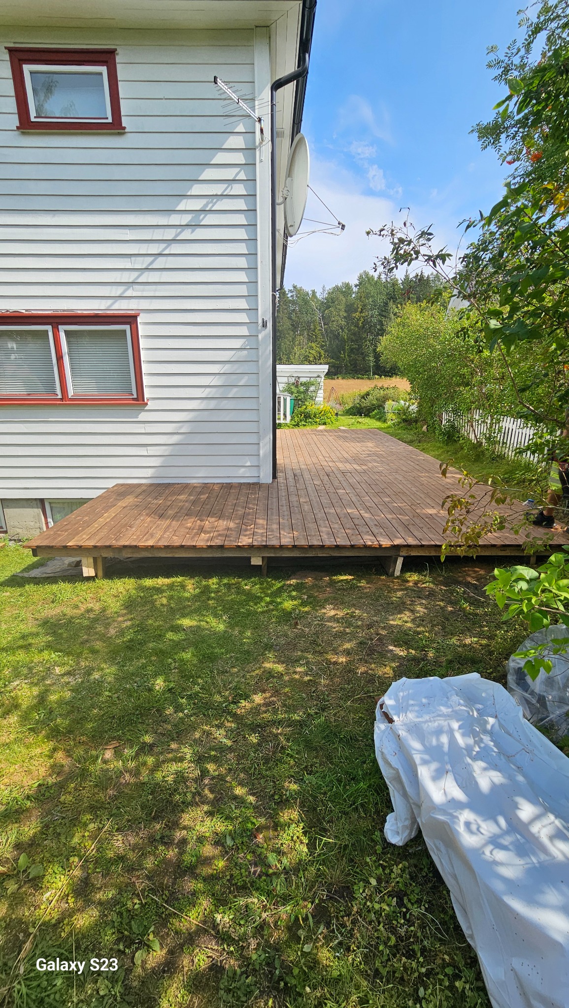 Nowy drewniany taras przylegający do białego domu z czerwonymi ramami okiennymi, widoczny trawnik i częściowo zakryty materiałem element wyposażenia.