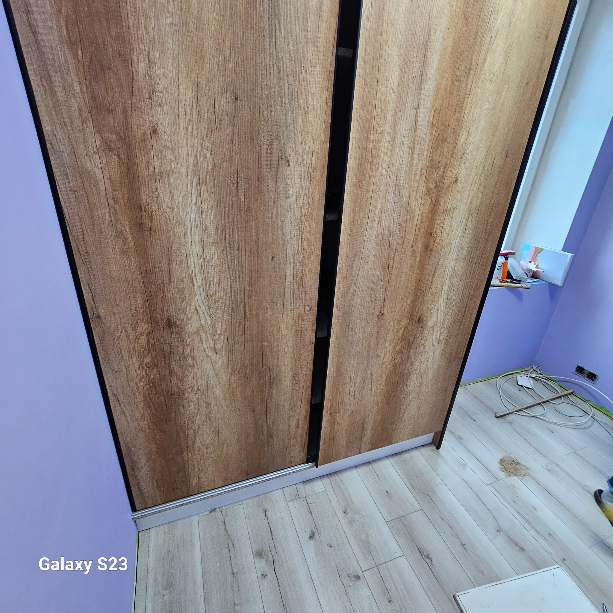 Montaż szafy przesuwnej z okleiną drewnopodobną w pokoju z fioletowymi ścianami i panelami podłogowymi, widoczne narzędzia i akcesoria montażowe.