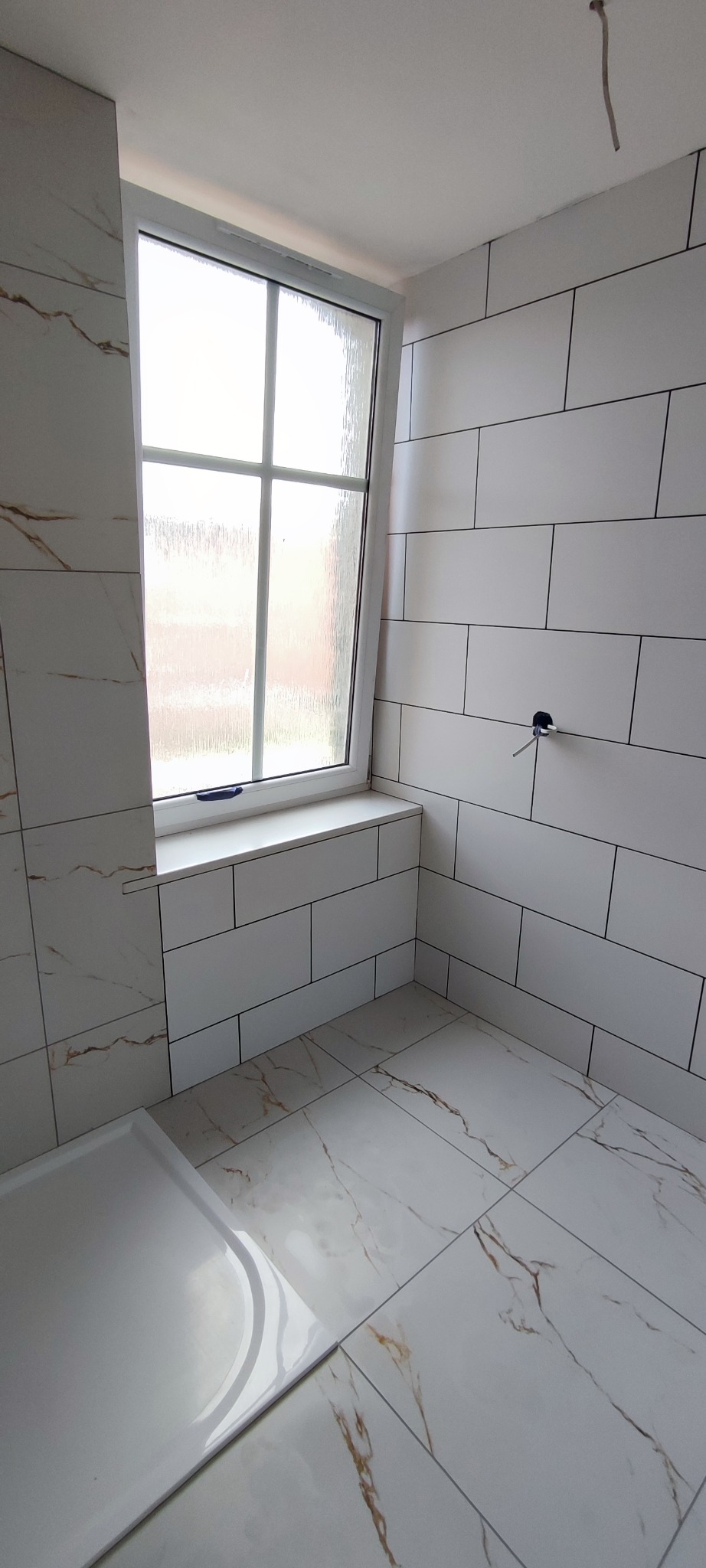 Biała łazienka z oknem, wykończona płytkami imitującymi marmur, z widocznym brodzikiem i wystającym przewodem elektrycznym, w trakcie prac wykończeniowych.