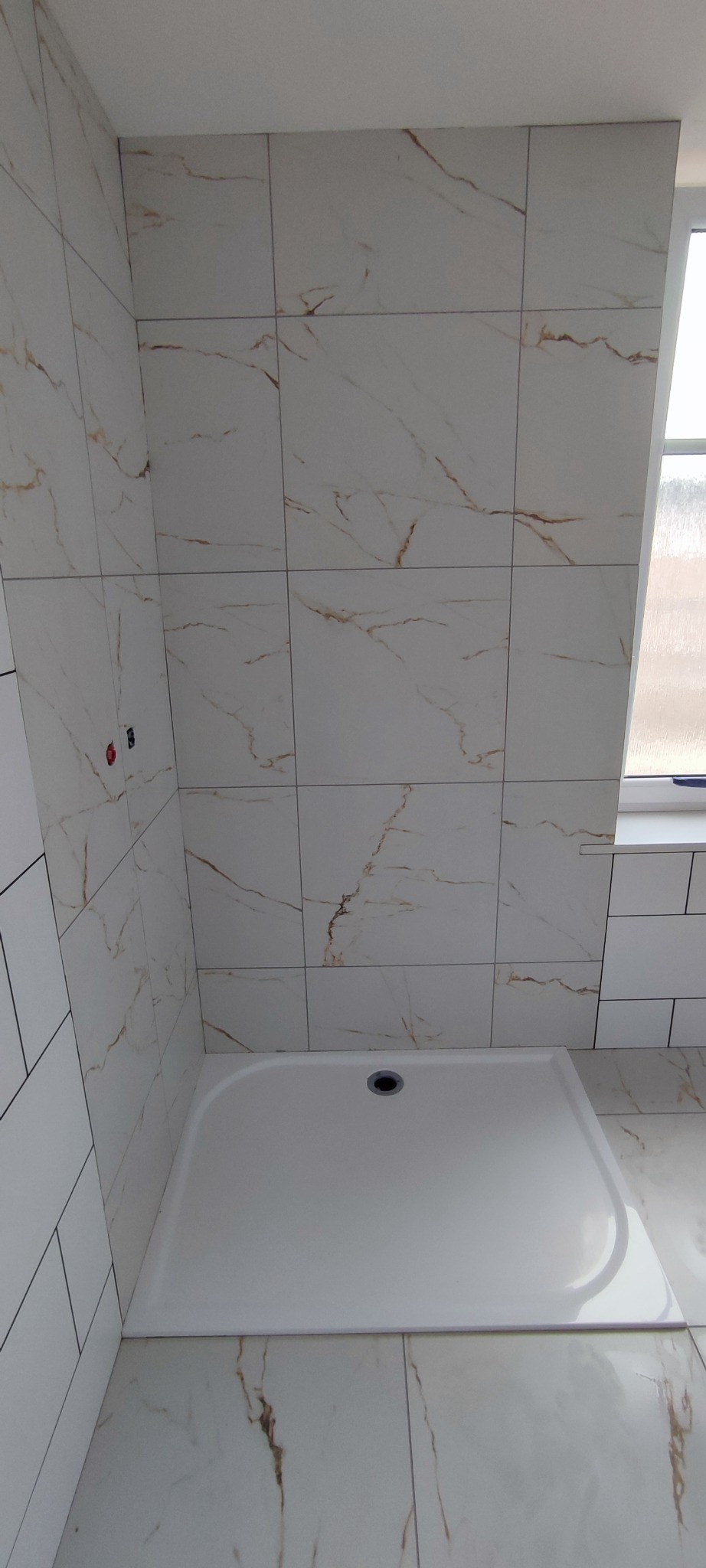 Nowa kabina prysznicowa z białym brodzikiem i płytkami imitującymi marmur na ścianach, widoczne fragmenty instalacji wodnej.