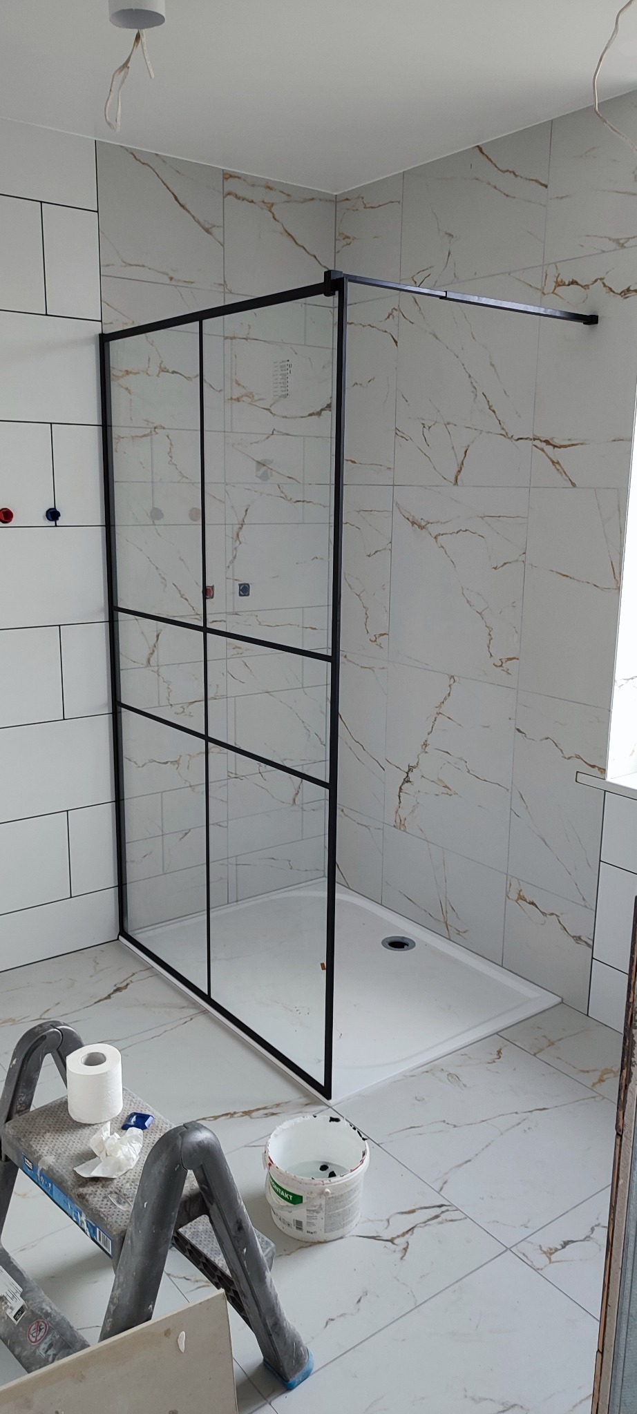 Nowoczesna kabina prysznicowa z czarnymi profilami i szklanymi ścianami, zamontowana w łazience wykończonej białymi płytkami imitującymi marmur, widoczna drabina malarska z rolką papieru...