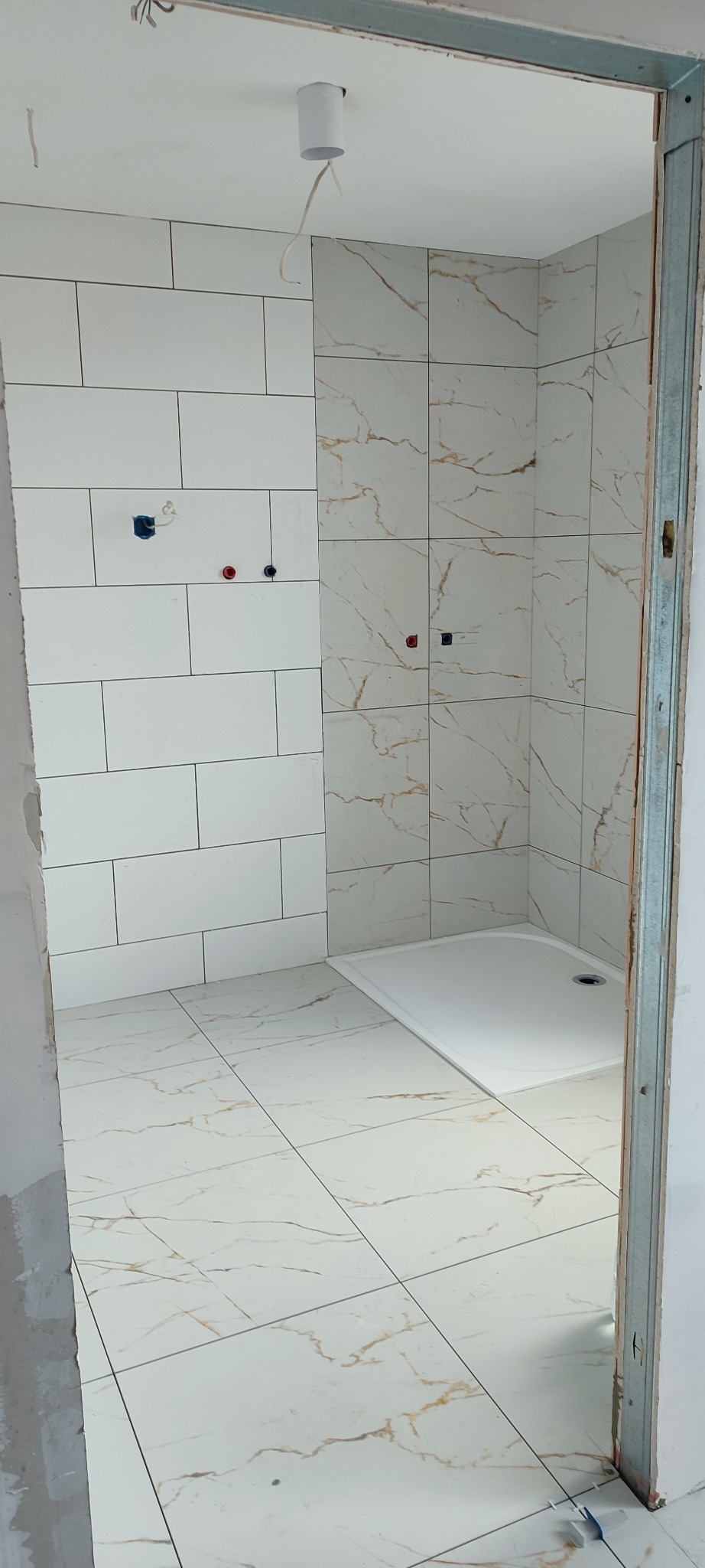 Wnętrze łazienki w trakcie układania płytek, widoczny brodzik i częściowo wykończone ściany z białych płytek o różnych wzorach, instalacja elektryczna w suficie i metalowa ościeżnica drzwi.