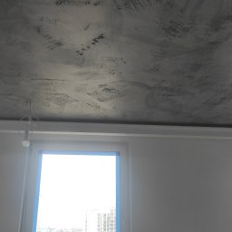 Dzmitry Sharakhouski - Szary sufit z efektem dekoracyjnym, widoczne ślady pędzla i nierównomierna faktura, okno z niebieską taśmą malarską, lampa wisząca na kablu, fragment rusztowania malarskiego po lewej stronie.