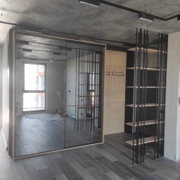 Dzmitry Sharakhouski - Nowoczesne wnętrze z przesuwną szafą z lustrzanymi drzwiami, drewnianą ścianką z wieszakami i metalowym regałem na tle surowego, betonowego sufitu z widocznym oświetleniem szynowym.