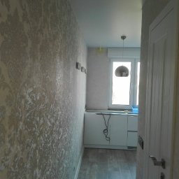 Dzmitry Sharakhouski - Wąski korytarz z dekoracyjną tapetą w beżowe ornamenty, drewnopodobna podłoga, biała szafka pod oknem i nowoczesna lampa wisząca.