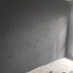 Dzmitry Sharakhouski - Ściana w szarym kolorze z efektem dekoracyjnym, widoczne puszki elektryczne, podłoga zabezpieczona folią malarską, pomieszczenie w trakcie remontu.