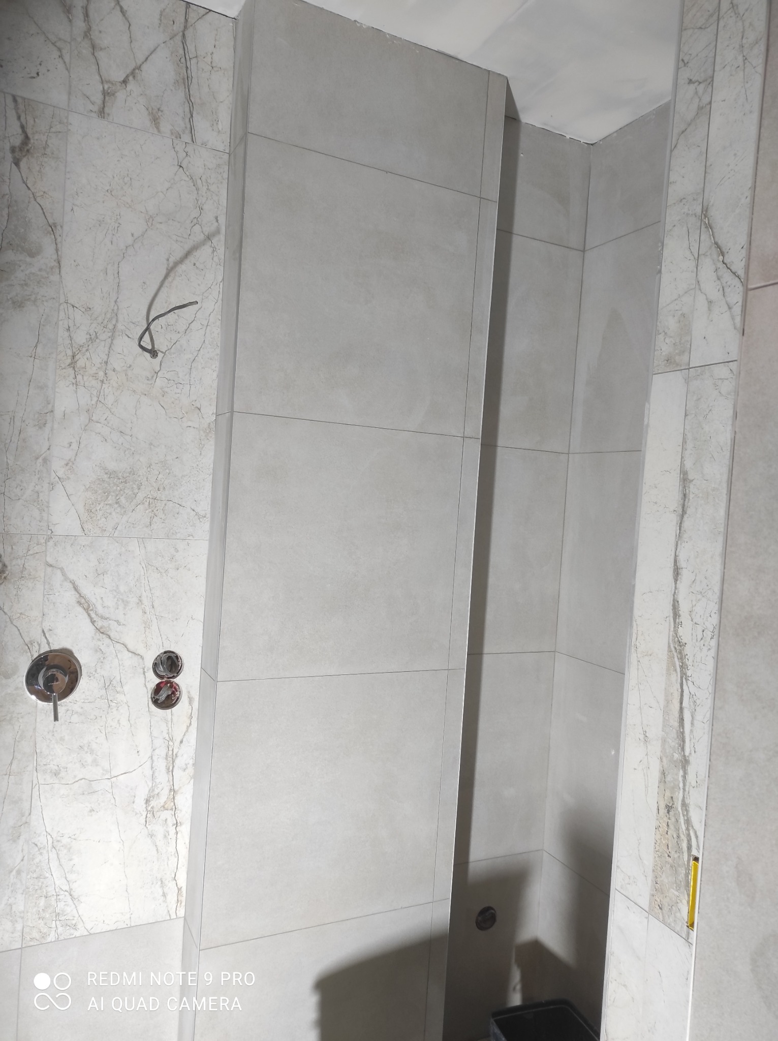 Wnętrze łazienki w trakcie wykańczania, z widocznymi płytkami imitującymi marmur na ścianie, szare płytki w strefie prysznicowej oraz wystające kable i armatura przed montażem.