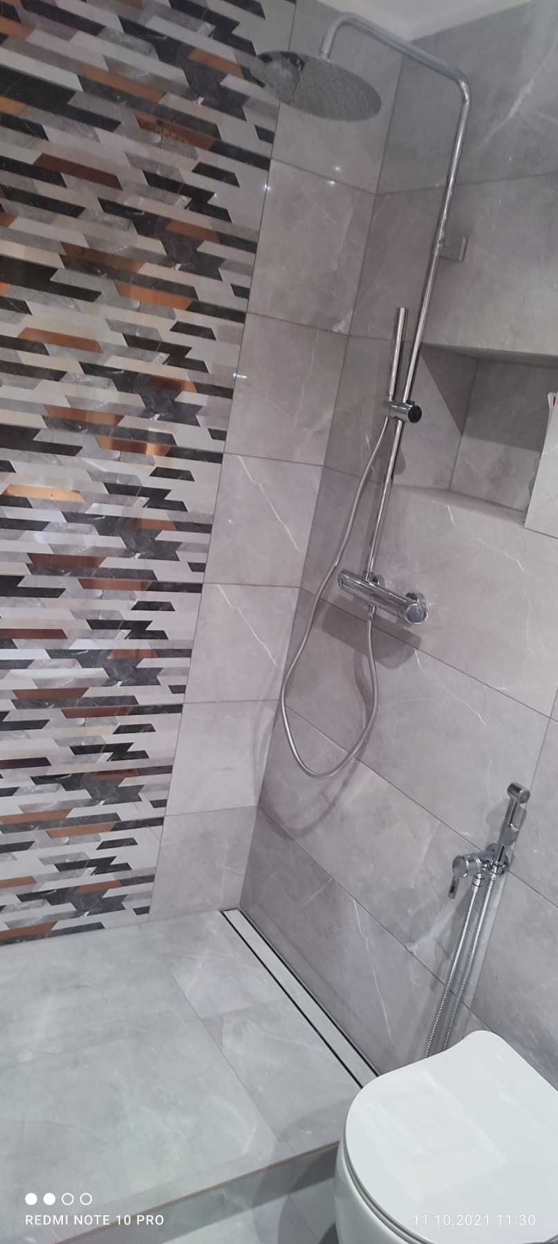 Nowoczesna łazienka z szarymi płytkami imitującymi marmur, kabina prysznicowa typu walk-in z odpływem liniowym i baterią podtynkową oraz toaleta wisząca.