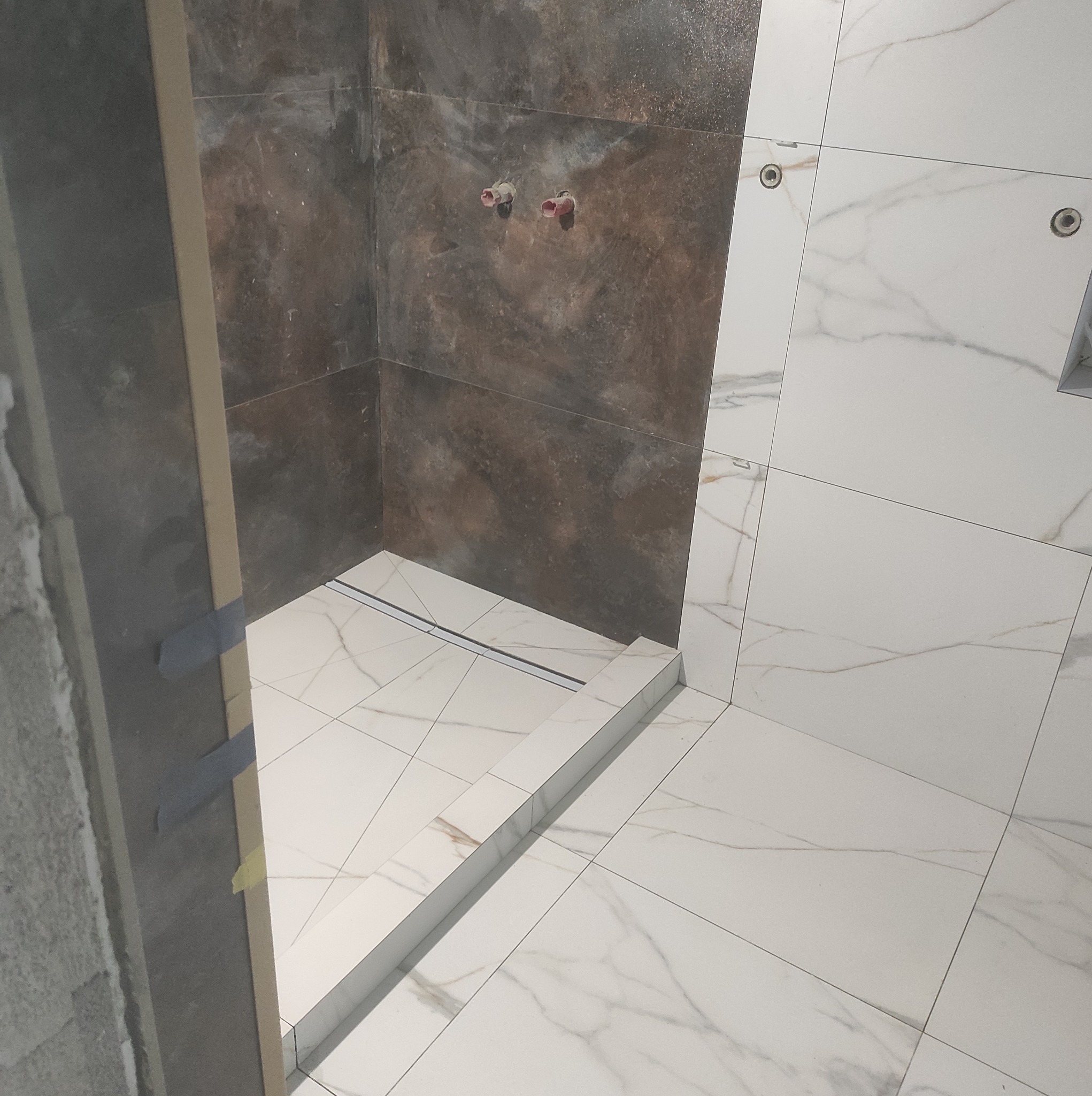 Wnętrze kabiny prysznicowej w trakcie wykańczania, widoczne brązowe i białe płytki imitujące marmur, odpływ liniowy oraz wystające rury instalacji wodnej.