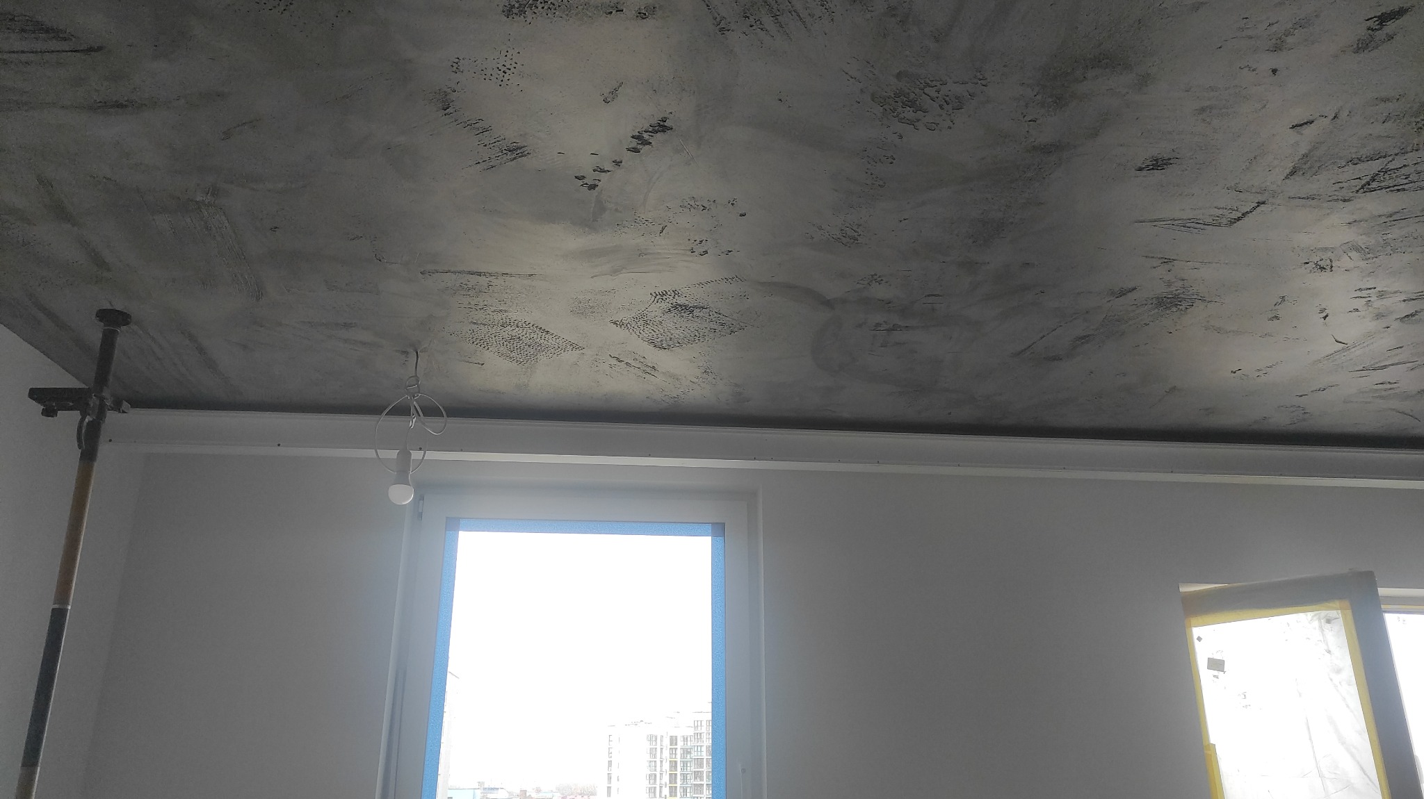 Szary sufit z efektem dekoracyjnym, widoczne ślady pędzla i nierównomierna faktura, okno z niebieską taśmą malarską, lampa wisząca na kablu, fragment rusztowania malarskiego po lewej stronie.