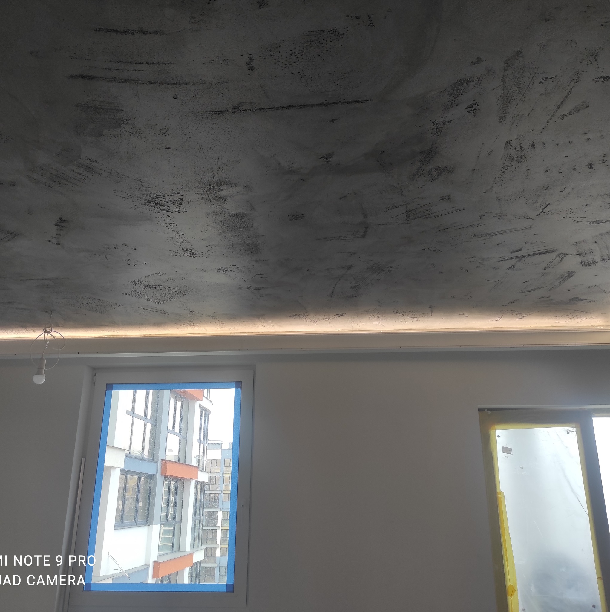 Szpachlowany sufit z efektem betonu, podświetlony listwą LED, okna zabezpieczone folią malarską, widok na budynek zza okna.