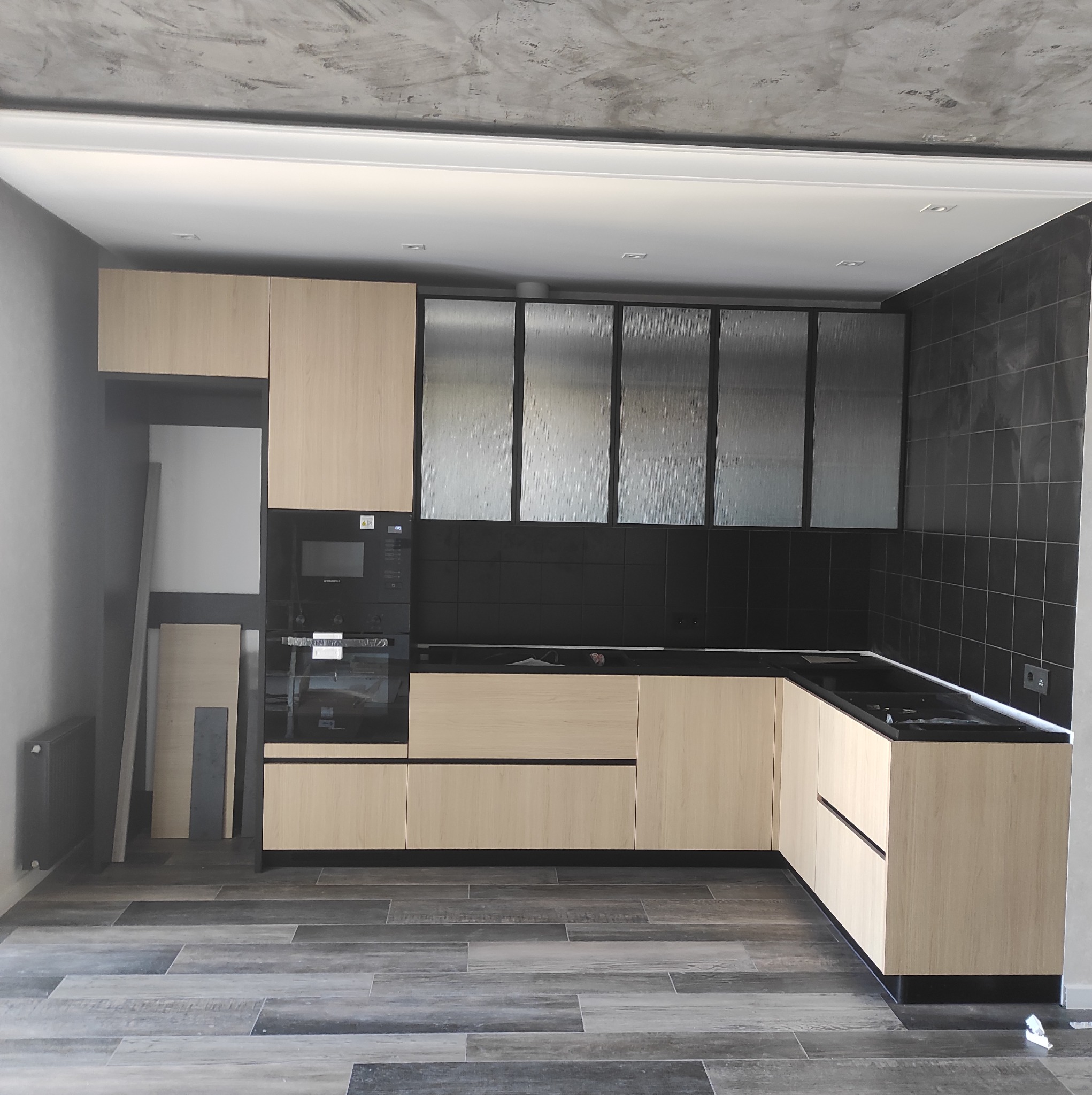 Nowoczesna kuchnia z jasnymi drewnianymi szafkami, czarnymi płytkami na ścianie i szklanymi szafkami wiszącymi z czarną ramą. Na podłodze szare płytki imitujące drewno. Ściany szare, sufit...