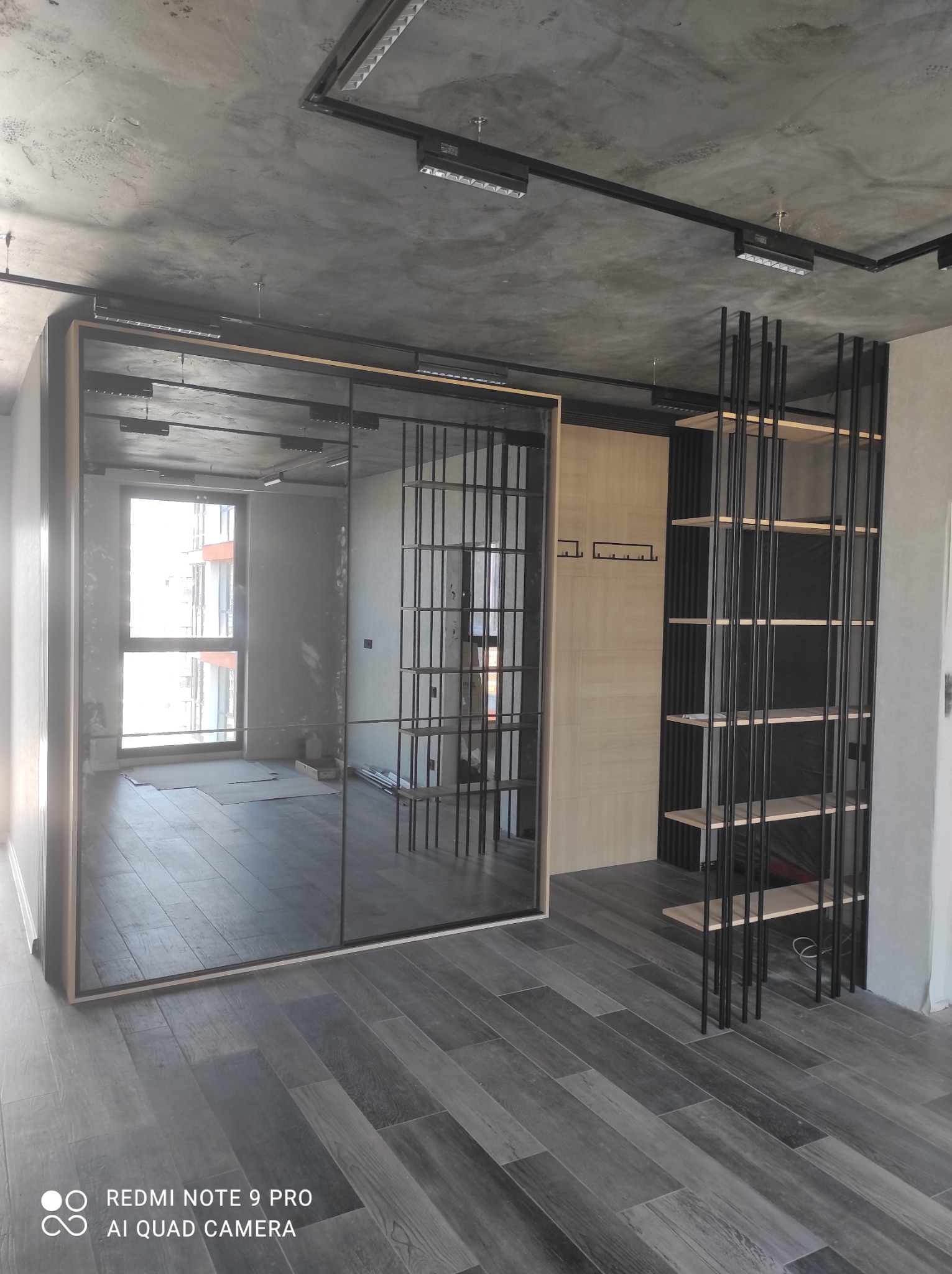 Nowoczesne wnętrze z przesuwną szafą z lustrzanymi drzwiami, drewnianą ścianką z wieszakami i metalowym regałem na tle surowego, betonowego sufitu z widocznym oświetleniem szynowym.