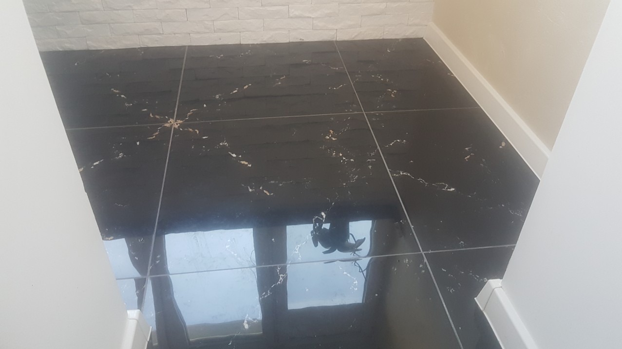 Błyszcząca, czarna podłoga z płytek imitujących marmur, ułożona w małym pomieszczeniu z białymi ścianami i listwami przypodłogowymi, z widocznym odbiciem okna w powierzchni płytek.