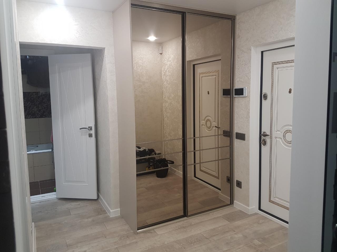 Jasne wnętrze przedpokoju z lustrzaną szafą przesuwną, widoczne drzwi wejściowe i wejście do łazienki z beżowymi płytkami.