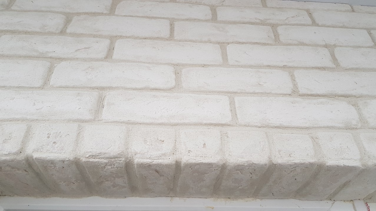Biała ściana z cegieł dekoracyjnych z wyraźną fakturą i szarawymi spoinami, ujęcie z dołu, widoczny fragment parapetu.