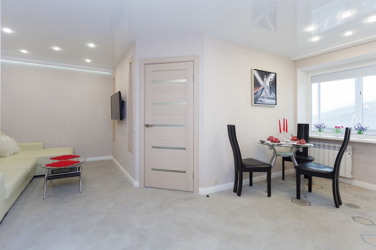 Jasne wnętrze mieszkania po remoncie z błyszczącym sufitem, beżowymi ścianami, szarą podłogą w heksagony, nowoczesnymi drzwiami z przeszkleniami, telewizorem na ścianie i stołem z krzesłami.