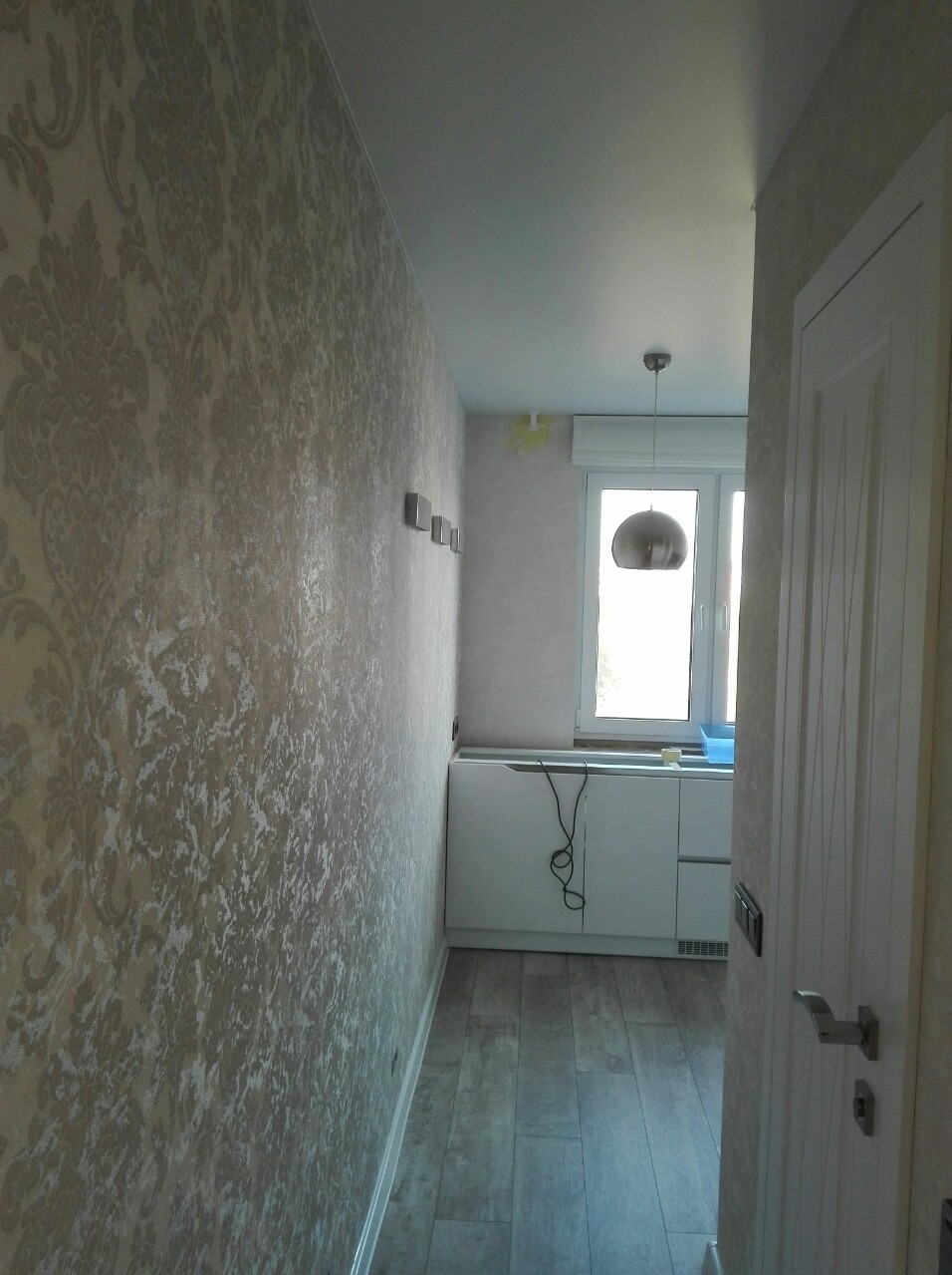 Wąski korytarz z dekoracyjną tapetą w beżowe ornamenty, drewnopodobna podłoga, biała szafka pod oknem i nowoczesna lampa wisząca.