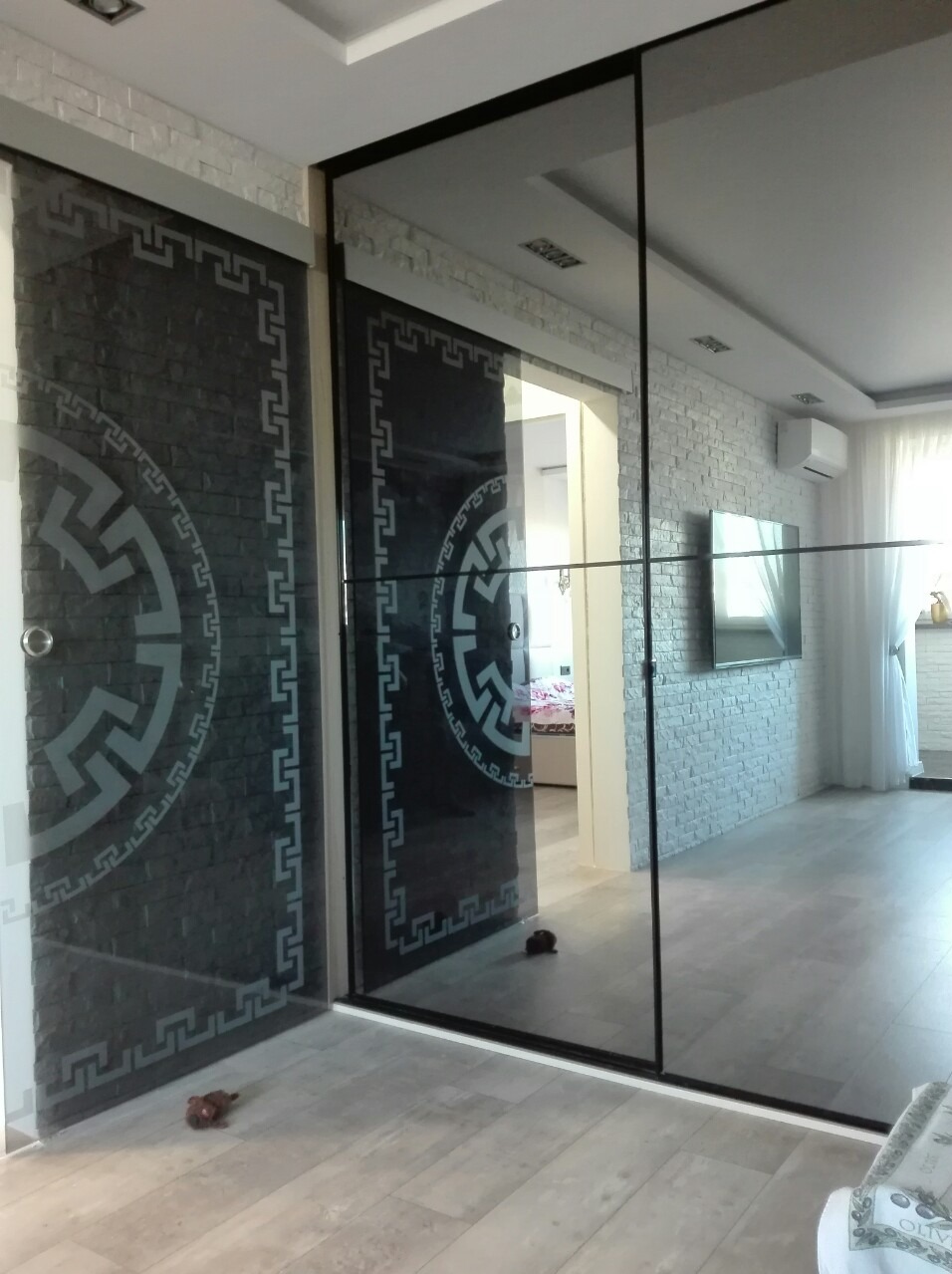 Nowoczesna szafa wnękowa z przesuwnymi drzwiami z dekoracyjnym wzorem na szkle, w tle ściana wykończona dekoracyjną cegłą i widok na wnętrze mieszkania.