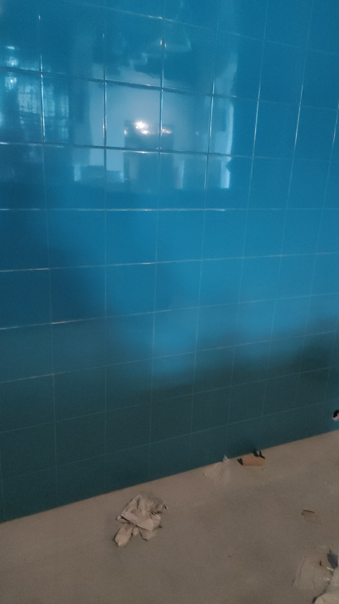 Ściana łazienki wyłożona błyszczącymi, turkusowymi płytkami ceramicznymi, widoczne niedociągnięcia na fugach i zabrudzenia na podłodze, co sugeruje trwający proces.