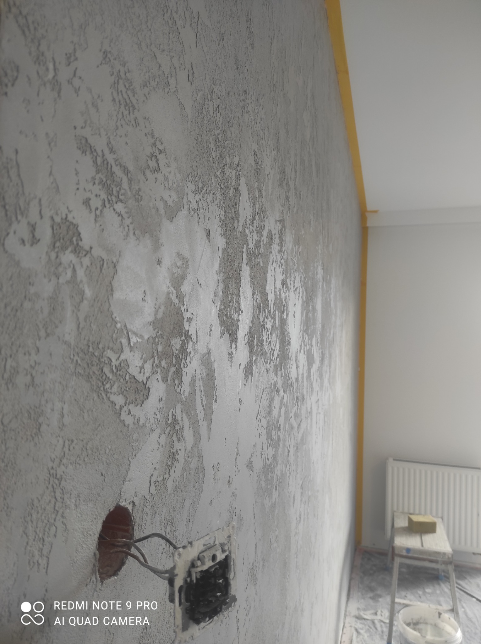 Ściana z szarą, dekoracyjną fakturą, zabezpieczona żółtą taśmą malarską przy suficie, z widocznym gniazdkiem elektrycznym w trakcie montażu i drabinką z gąbką w tle.