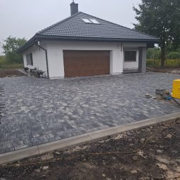 OL-BRUK OLEH TVERDOKHLIB - Nowoczesny dom jednorodzinny z szarą kostką brukową na podjeździe, widoczne obrzeża i przygotowane do ułożenia elementy, w tle zaparkowana taczka i agregat prądotwórczy.