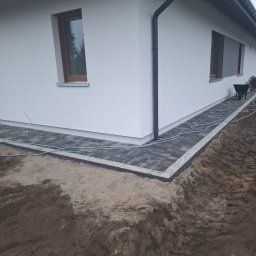 OL-BRUK OLEH TVERDOKHLIB - Świeżo ułożona ścieżka z grafitowej kostki brukowej z jasnoszarym obrzeżem okalająca narożnik domu z białym tynkiem, widoczny fragment ogrodu z ziemią, w tle taczka.