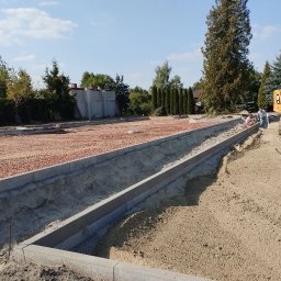 OL-BRUK OLEH TVERDOKHLIB - Przygotowanie podłoża pod chodnik z obrzeżami betonowymi, widoczny koparka i pracownicy w tle, teren wyrównany żwirem