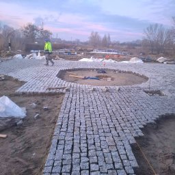OL-BRUK OLEH TVERDOKHLIB - Obszar w trakcie wykańczania z szarej kostki granitowej, widoczny pracownik w odblaskowej kurtce, w tle elementy budowy i pochmurne niebo.
