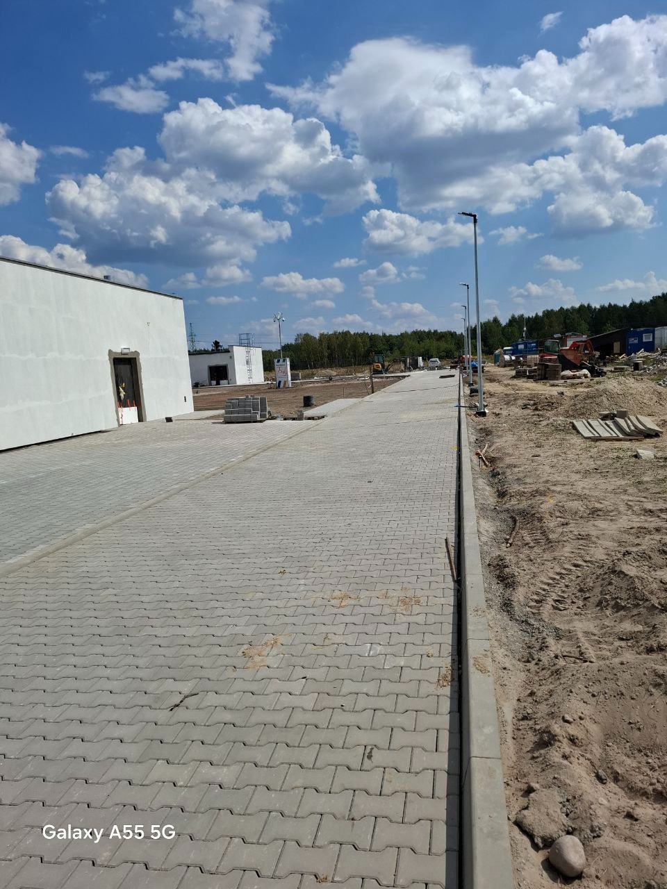 Długa droga wyłożona szarą kostką brukową typu 'I' w perspektywie, z widocznymi budynkami w tle i błękitnym niebem z chmurami.