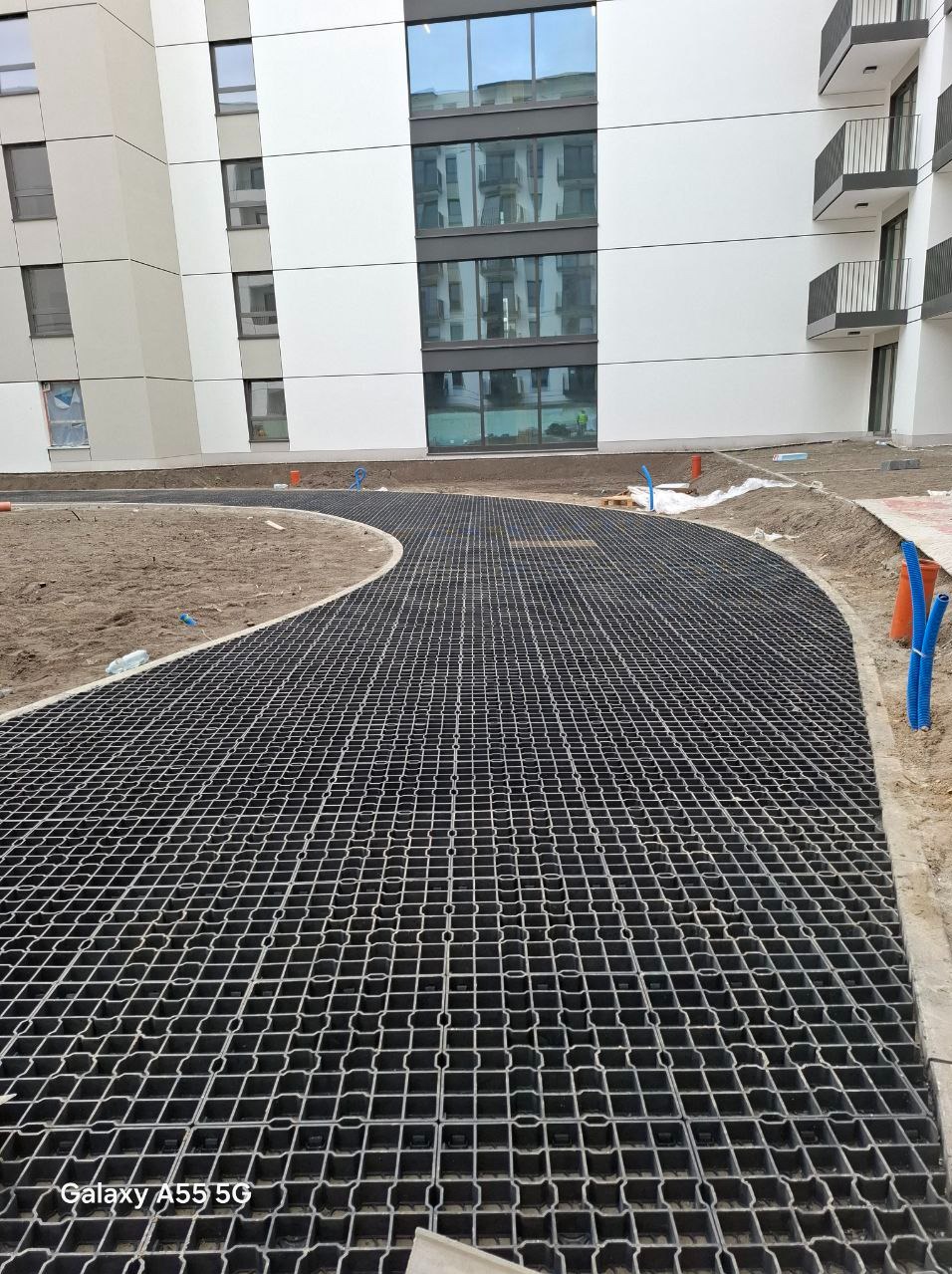 Układanie czarnej, plastikowej kratki trawnikowej na piasku wzdłuż zakrzywionej ścieżki przed nowoczesnym budynkiem mieszkalnym z widocznymi instalacjami wodnymi.