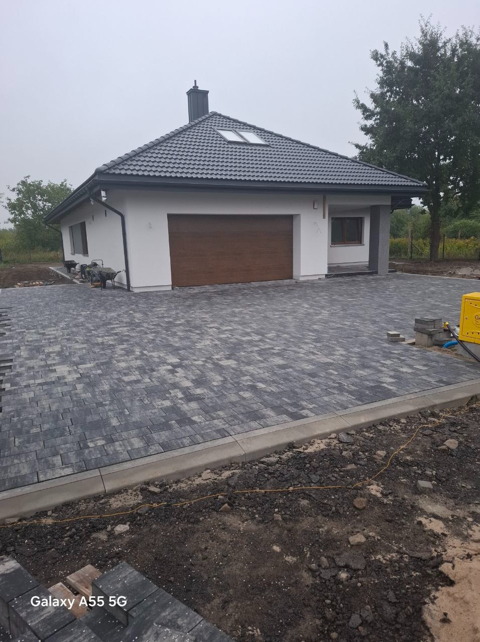 Nowoczesny dom jednorodzinny z szarą kostką brukową na podjeździe, widoczne obrzeża i przygotowane do ułożenia elementy, w tle zaparkowana taczka i agregat prądotwórczy.
