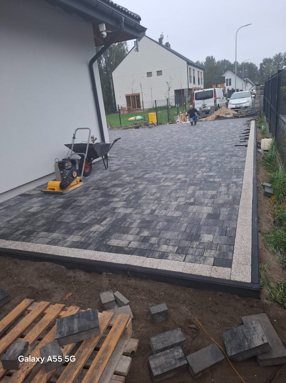 Ułożona nawierzchnia z kostki brukowej w odcieniach szarości z jasnym obramowaniem, widoczna zagęszczarka i taczka, w tle budynek mieszkalny i pracownik podczas realizacji.