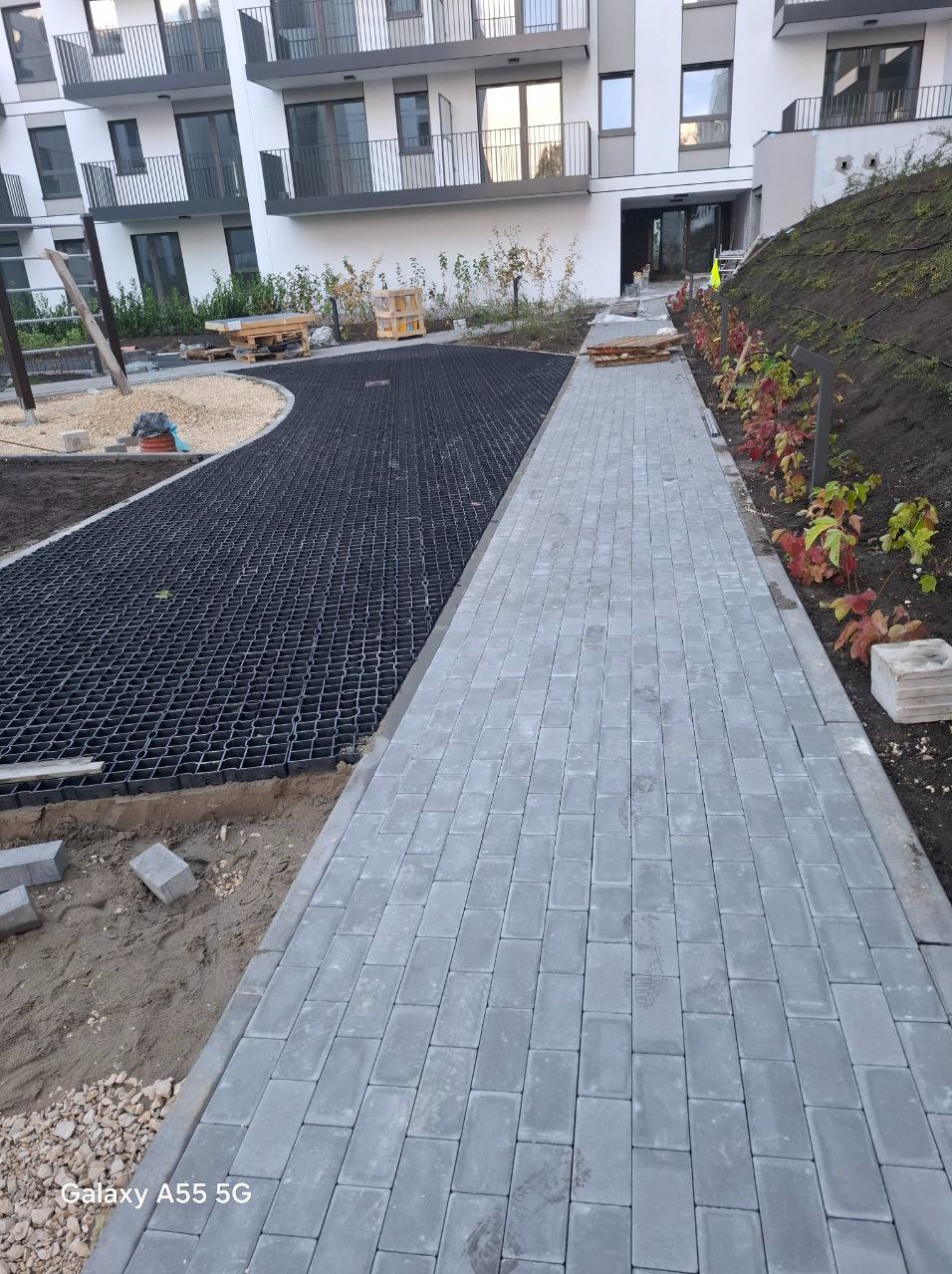 Ułożona ścieżka z szarej kostki brukowej prowadząca wzdłuż budynku mieszkalnego, obok fragmentu terenu z czarną geokratą i nasadzeniami roślin.