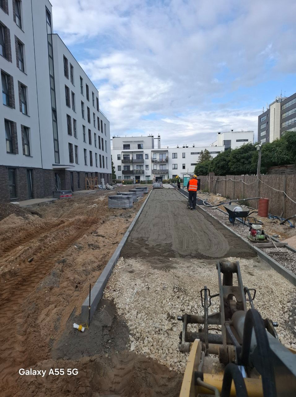 Prace ziemne w Warszawie: utwardzanie drogi dojazdowej na terenie budowy osiedla mieszkaniowego, widoczny ubijak wibracyjny i miniładowarka.