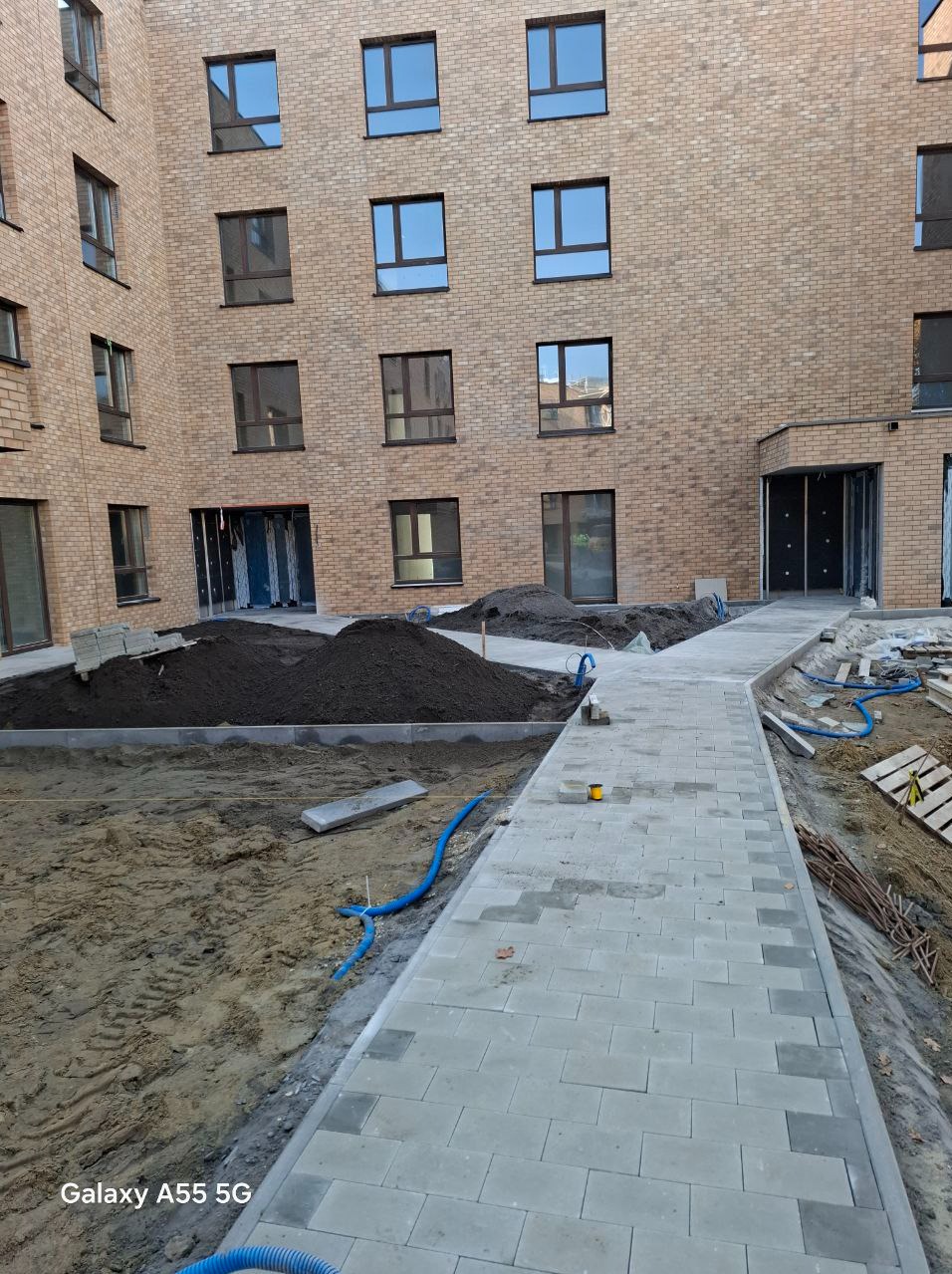 Układanie szarej kostki brukowej przed budynkiem z cegły, widoczne hałdy ziemi i niebieski wąż do nawadniania.