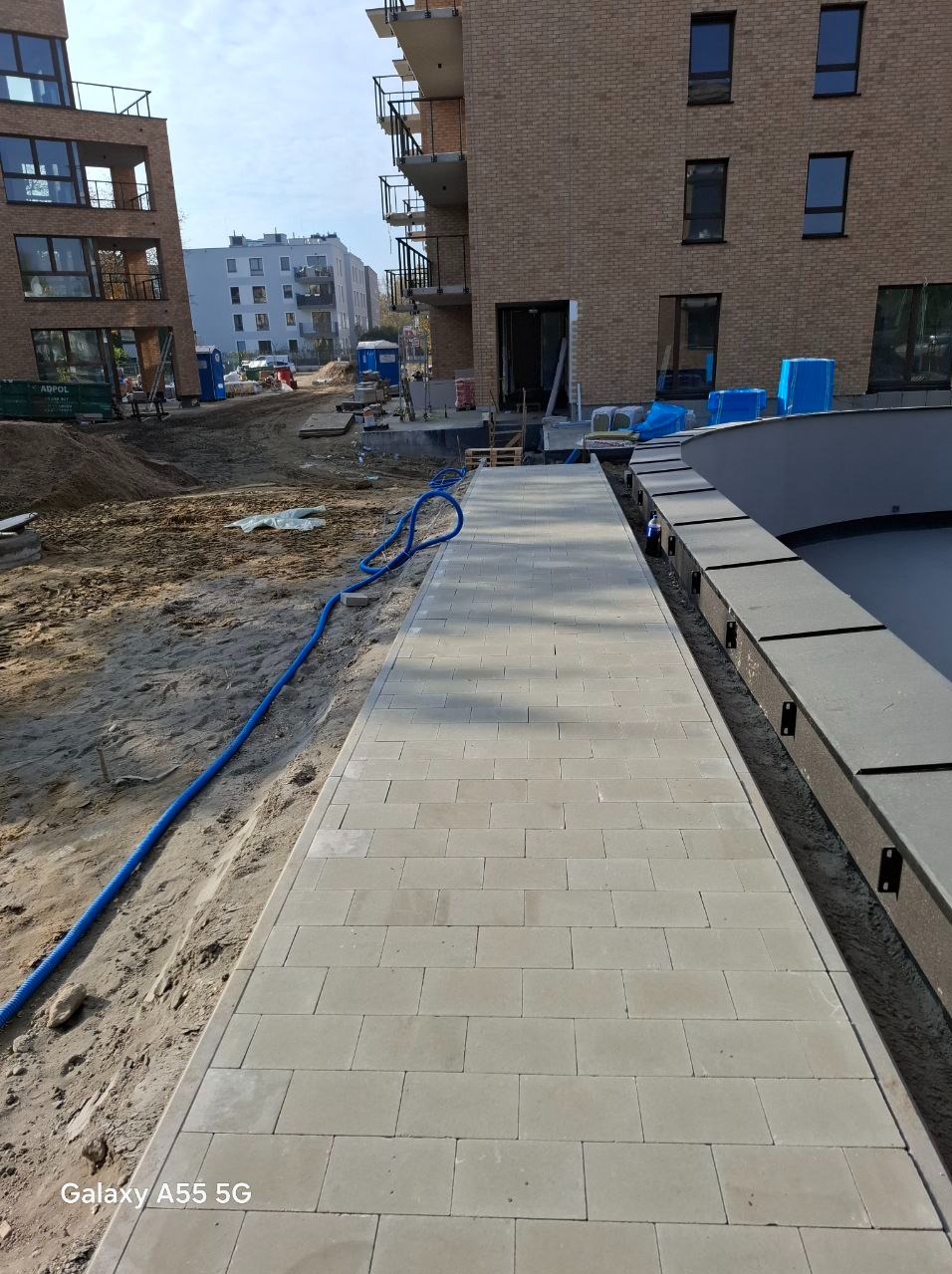 Świeżo ułożony chodnik z jasnoszarej kostki brukowej prowadzący wzdłuż budynku mieszkalnego w trakcie budowy, widoczny niebieski wąż i przygotowane podłoże.
